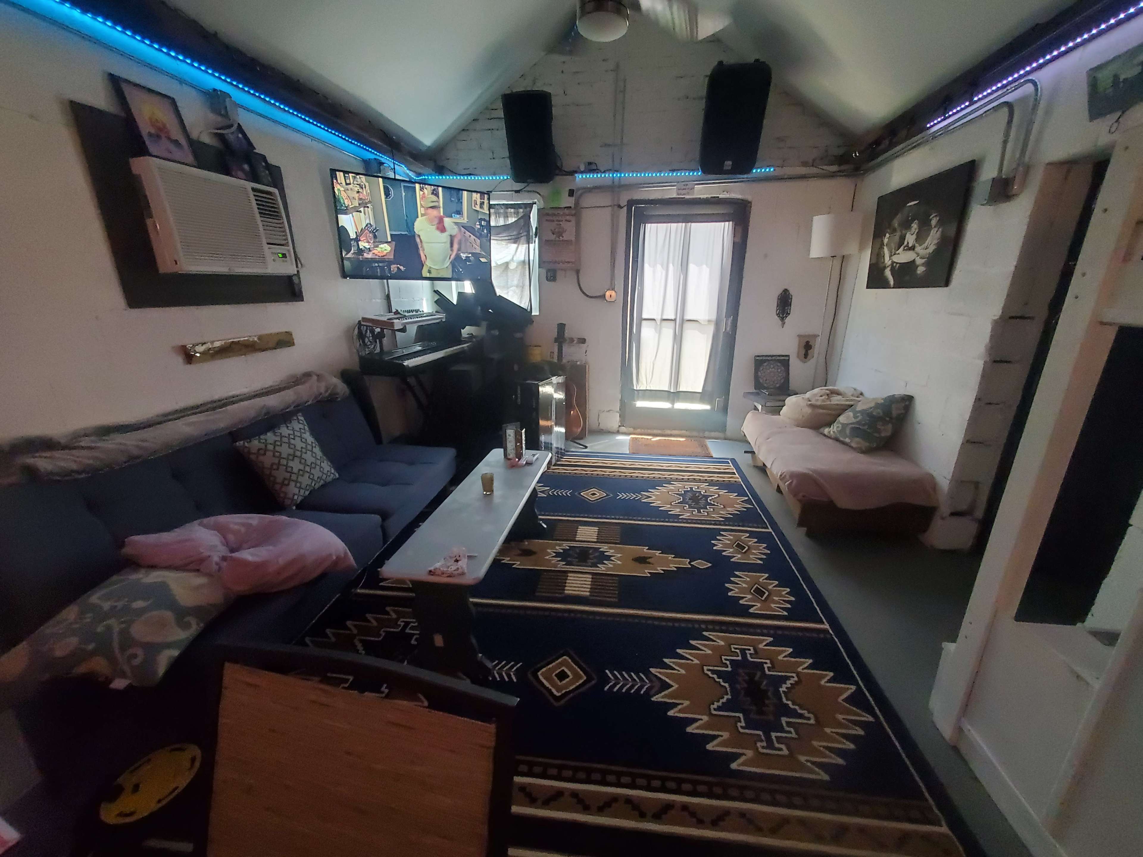 The image shows a cozy interior space with a blue couch, a coffee table, a patterned rug, and a door leading to a balcony, illuminated by ambient lighting.