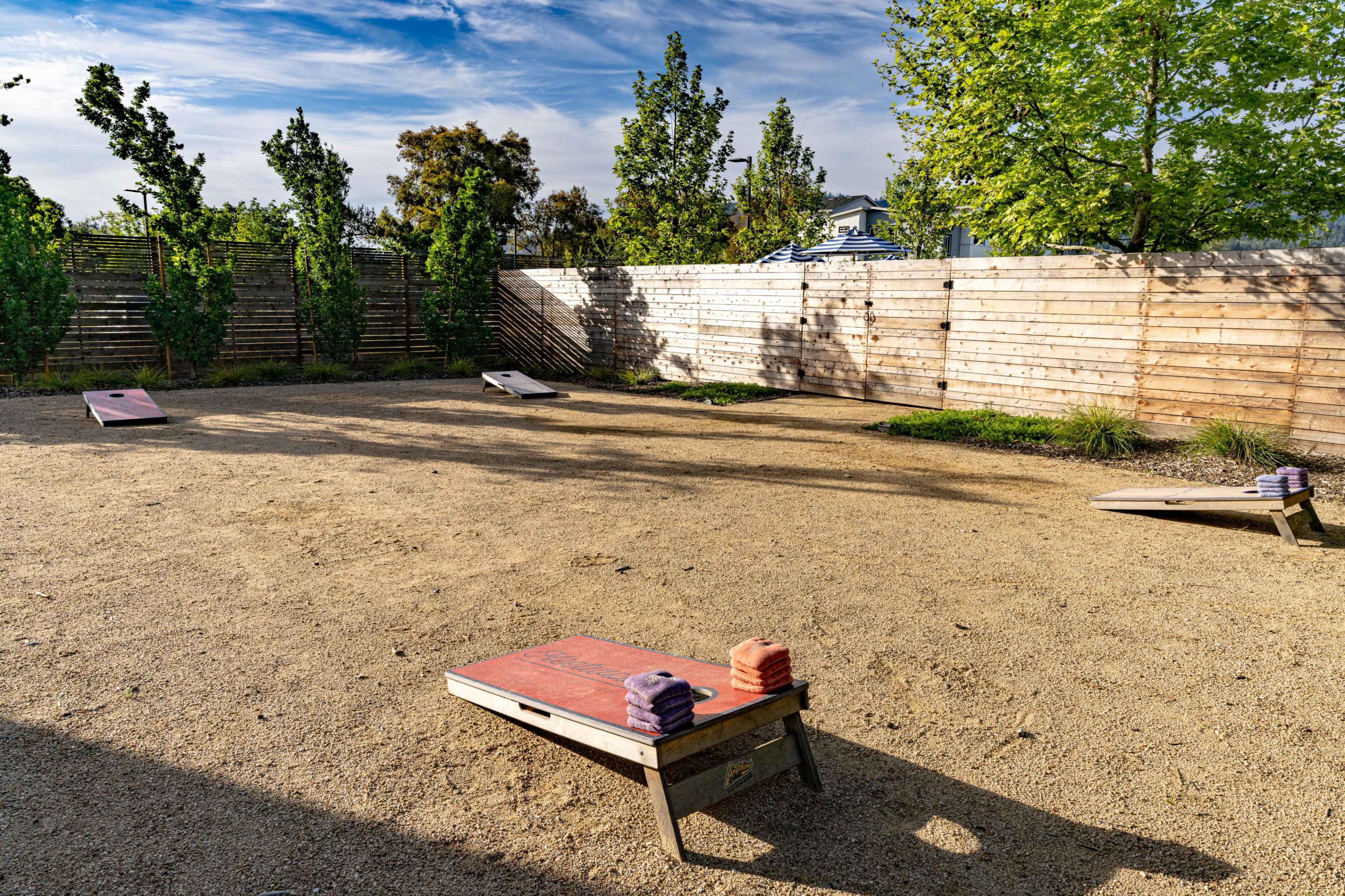 The image shows a sandy outdoor space with two elevated wooden platforms for cornhole and a lined fence with trees in the background.