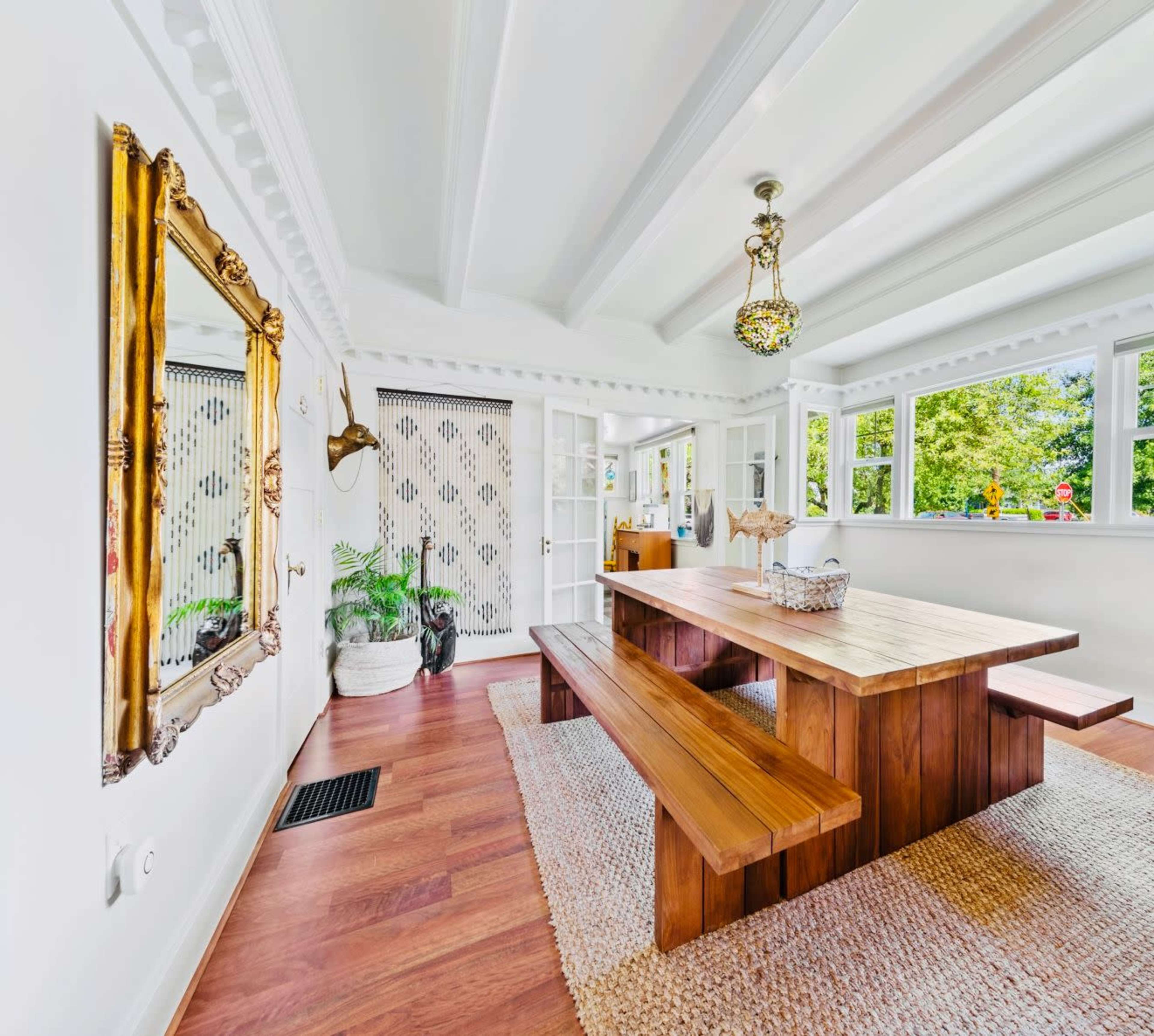 A bright dining area featuring a large wooden table with bench seating, surrounded by windows and decorated with a mirror and plants.
