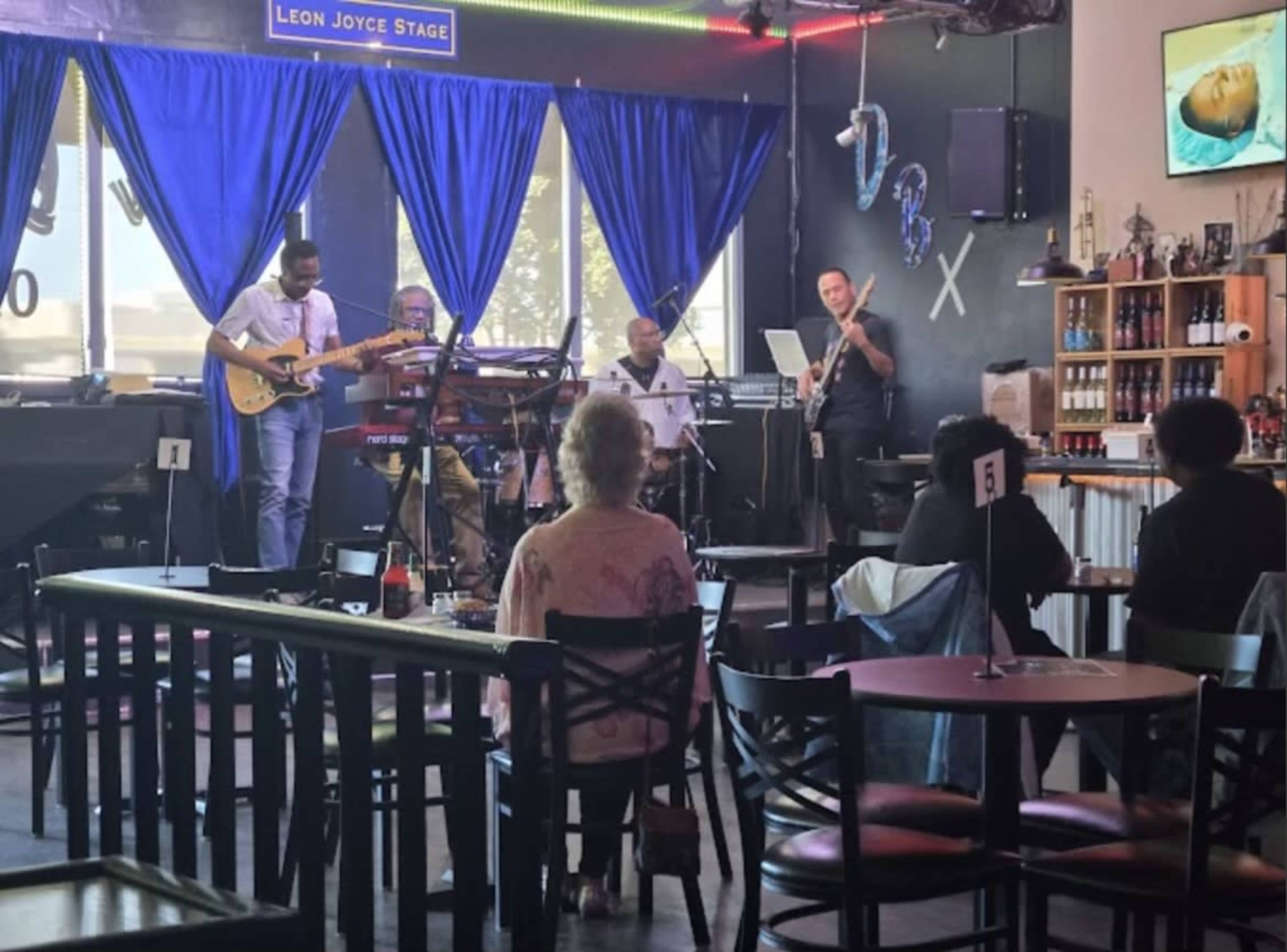 A small band performs on stage in a dimly lit venue with blue curtains, while an audience sits at tables in the foreground.