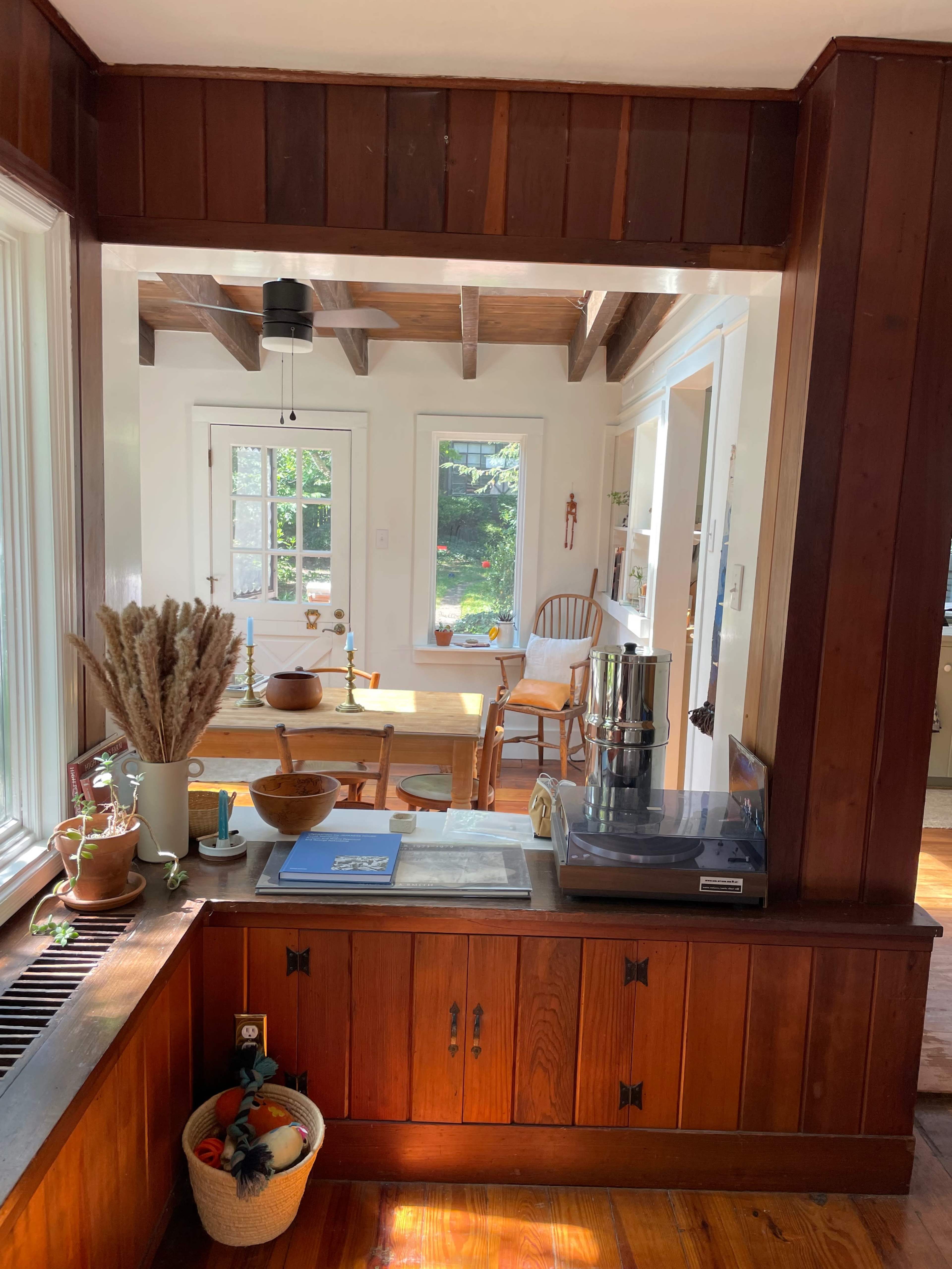 A wooden kitchen area with a view into a dining space featuring a table and chairs, illuminated by natural light through windows.