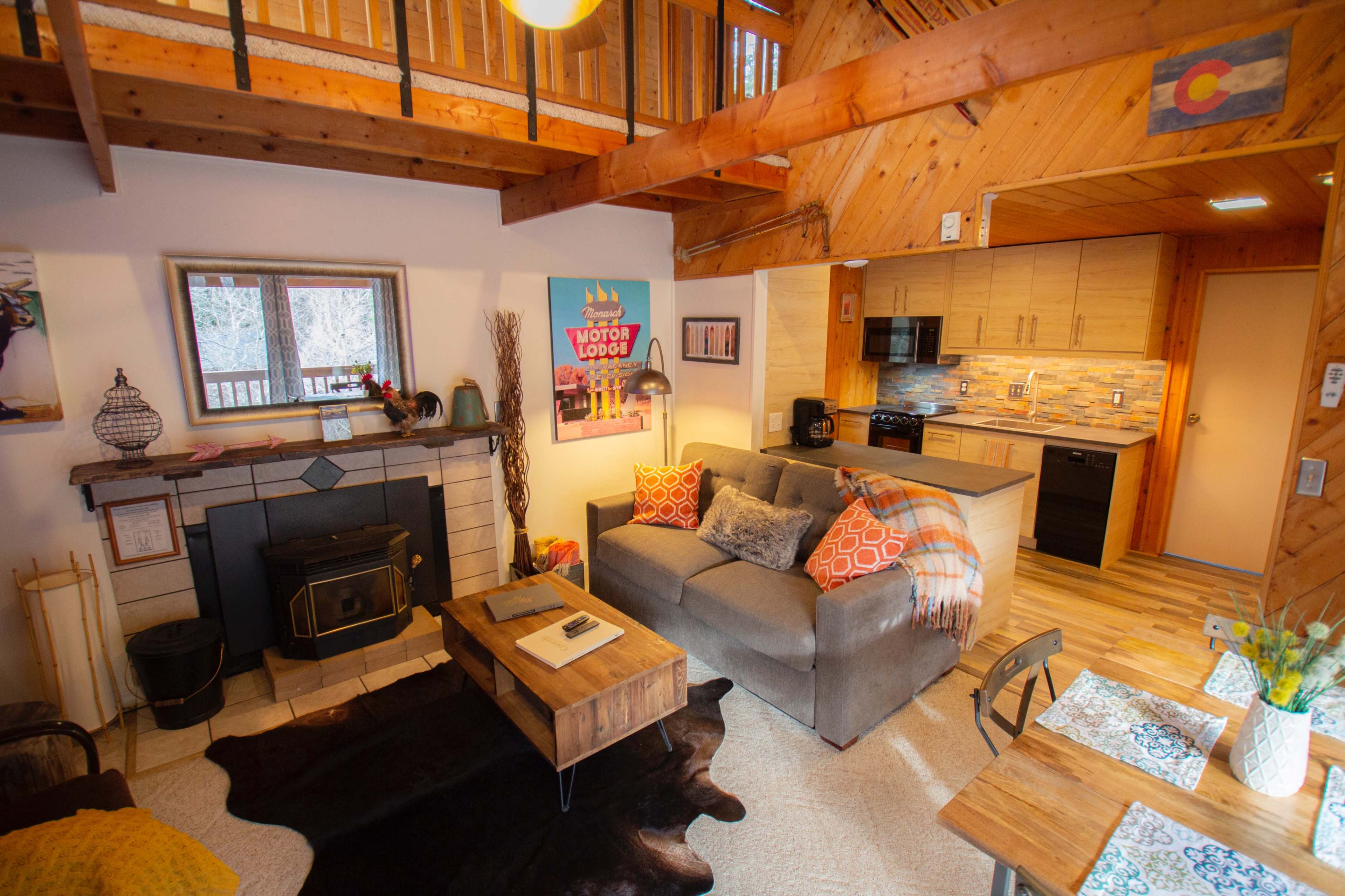 A cozy cabin interior featuring a living area with a sofa, a coffee table, and a fireplace, alongside a kitchen and dining space.