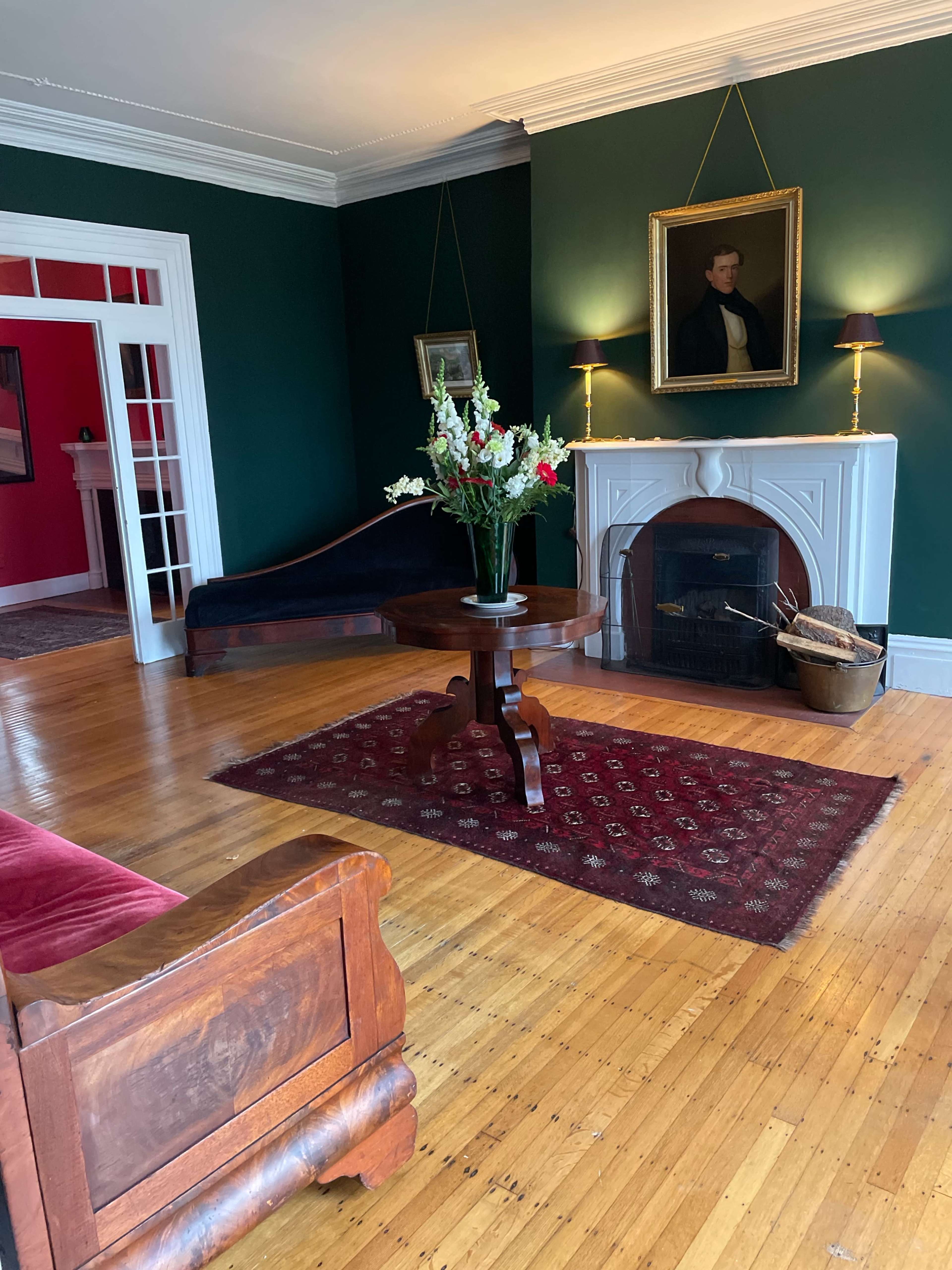 The image shows a room with dark green walls, a wooden floor, a fireplace with a portrait above it, a round table with a floral arrangement, and a sofa against the wall.
