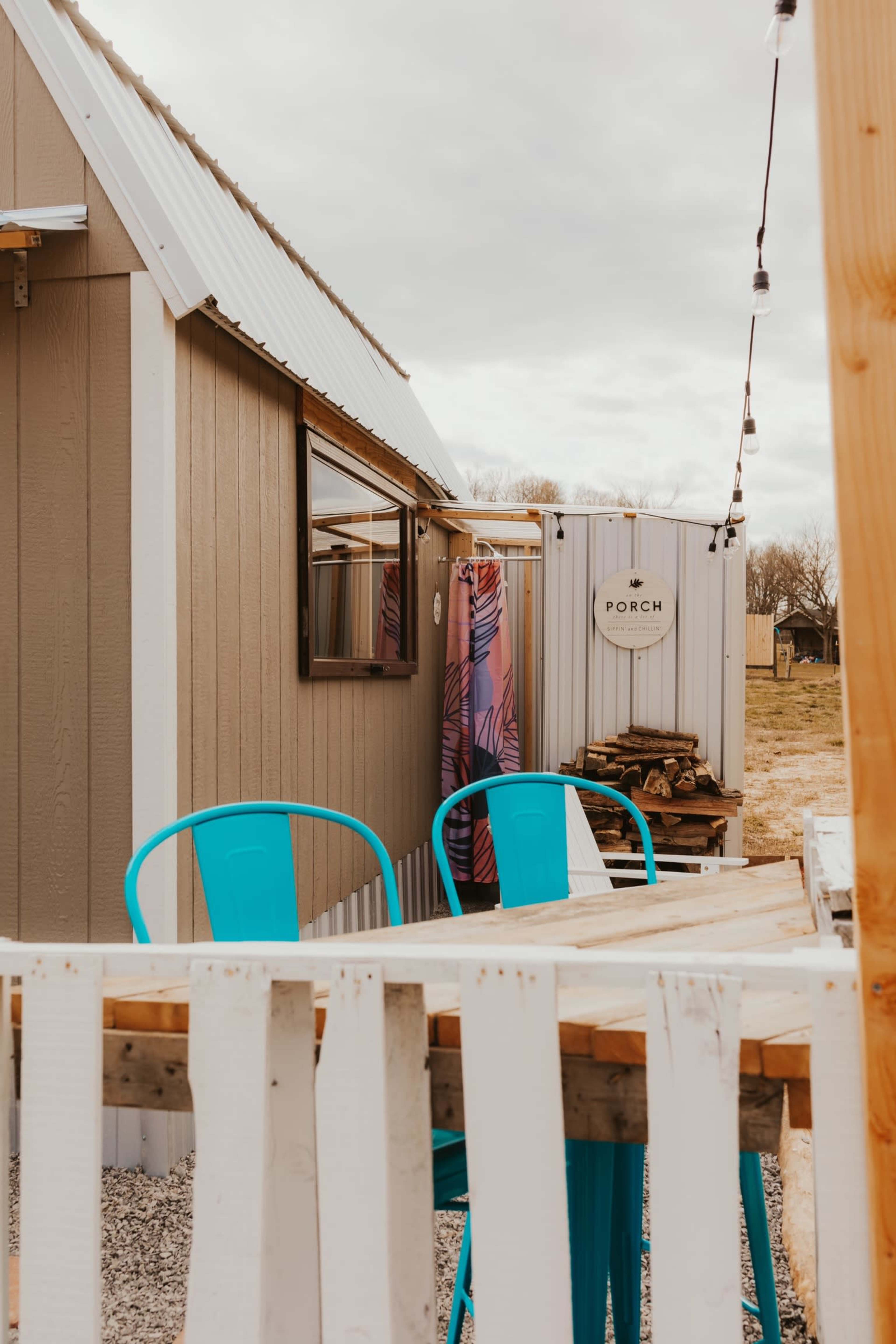 A small cabin with a light-colored exterior features a wooden table and chairs outside, and a nearby shed labeled "PORCH."