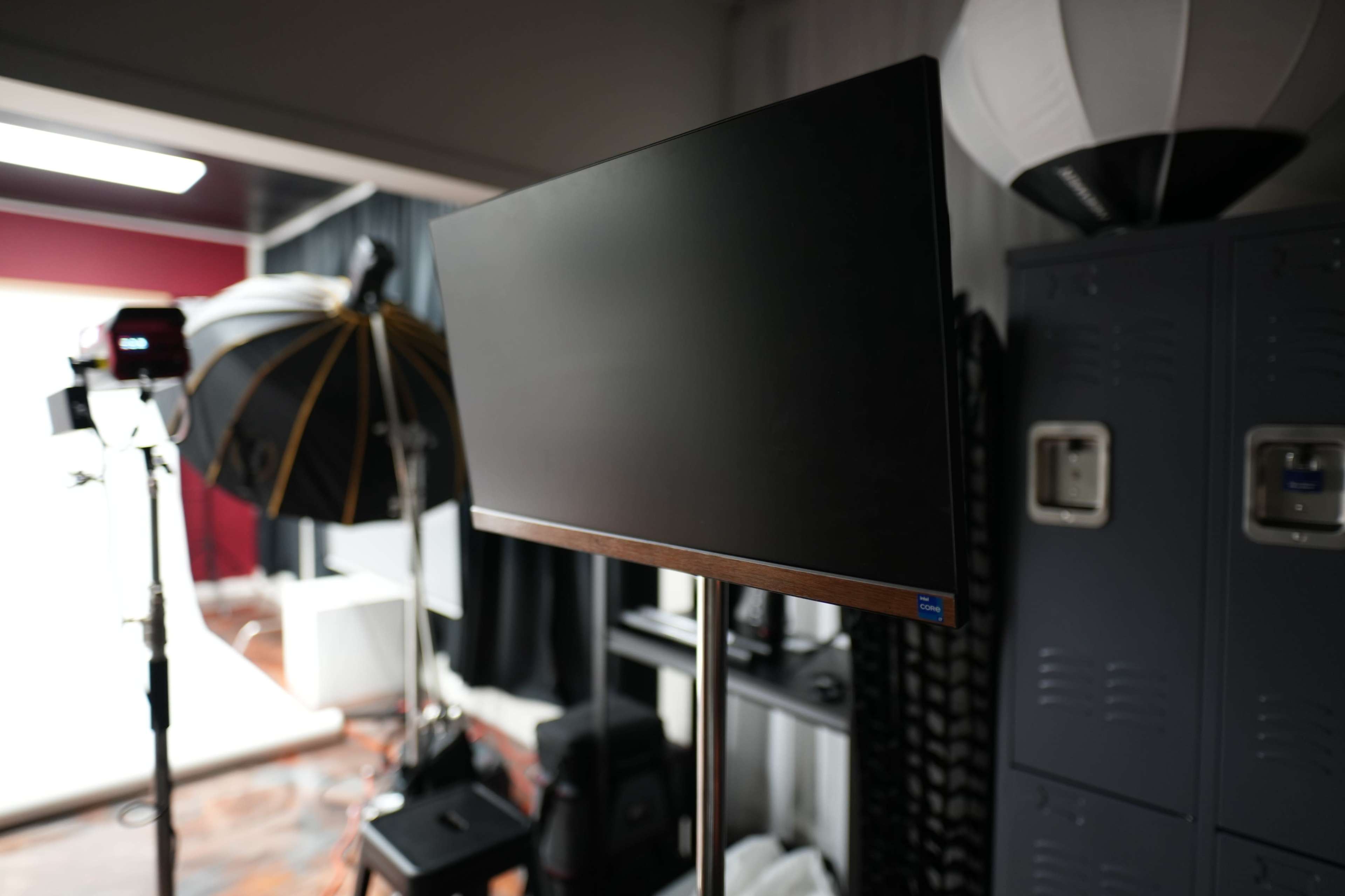 A flat-screen monitor is mounted on a stand in a studio setting, with various photography equipment and backdrops visible in the background.