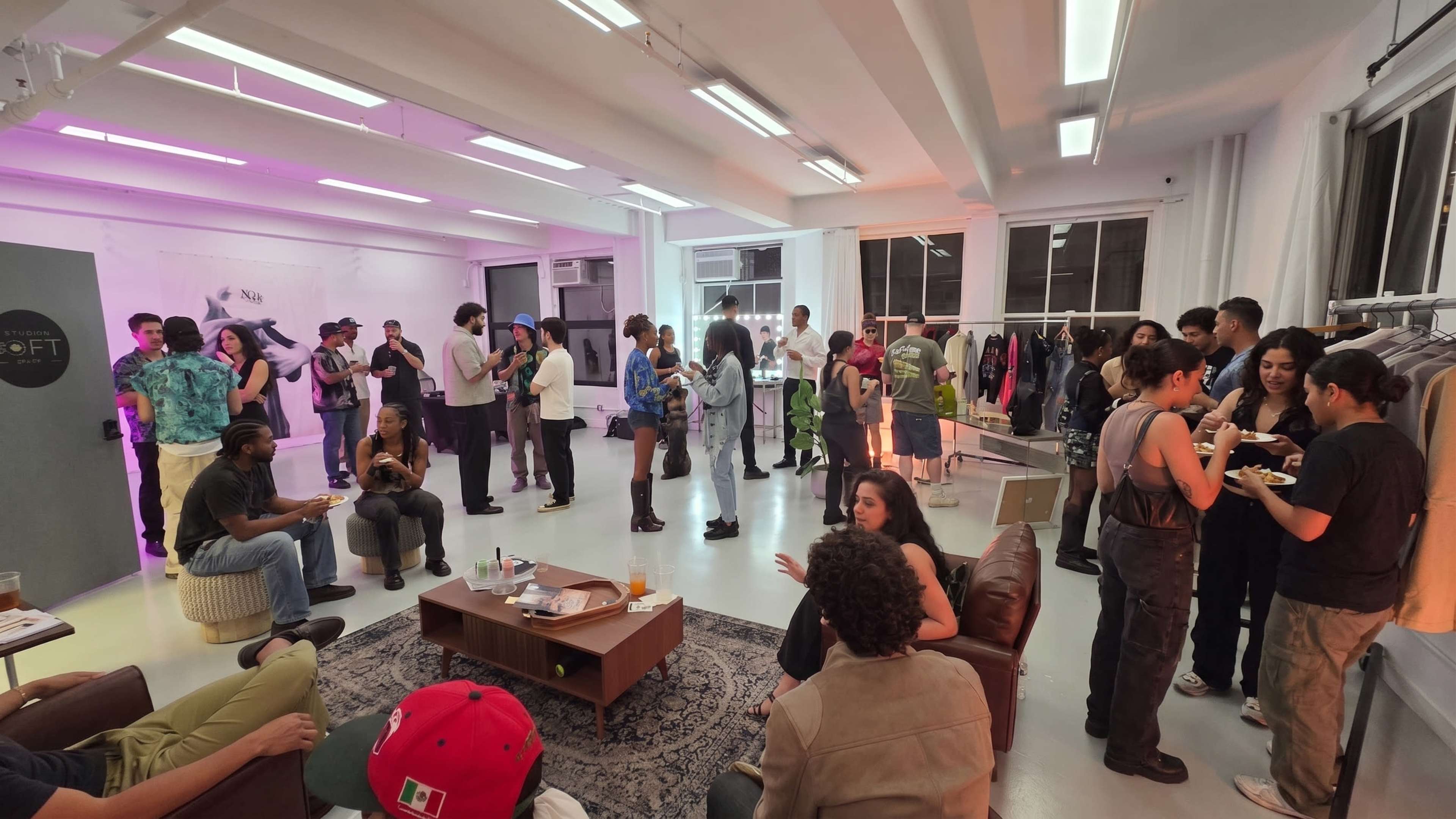 A large gathering takes place in a well-lit room with people chatting, standing, and sitting around furniture while food and drinks are accessible on tables.