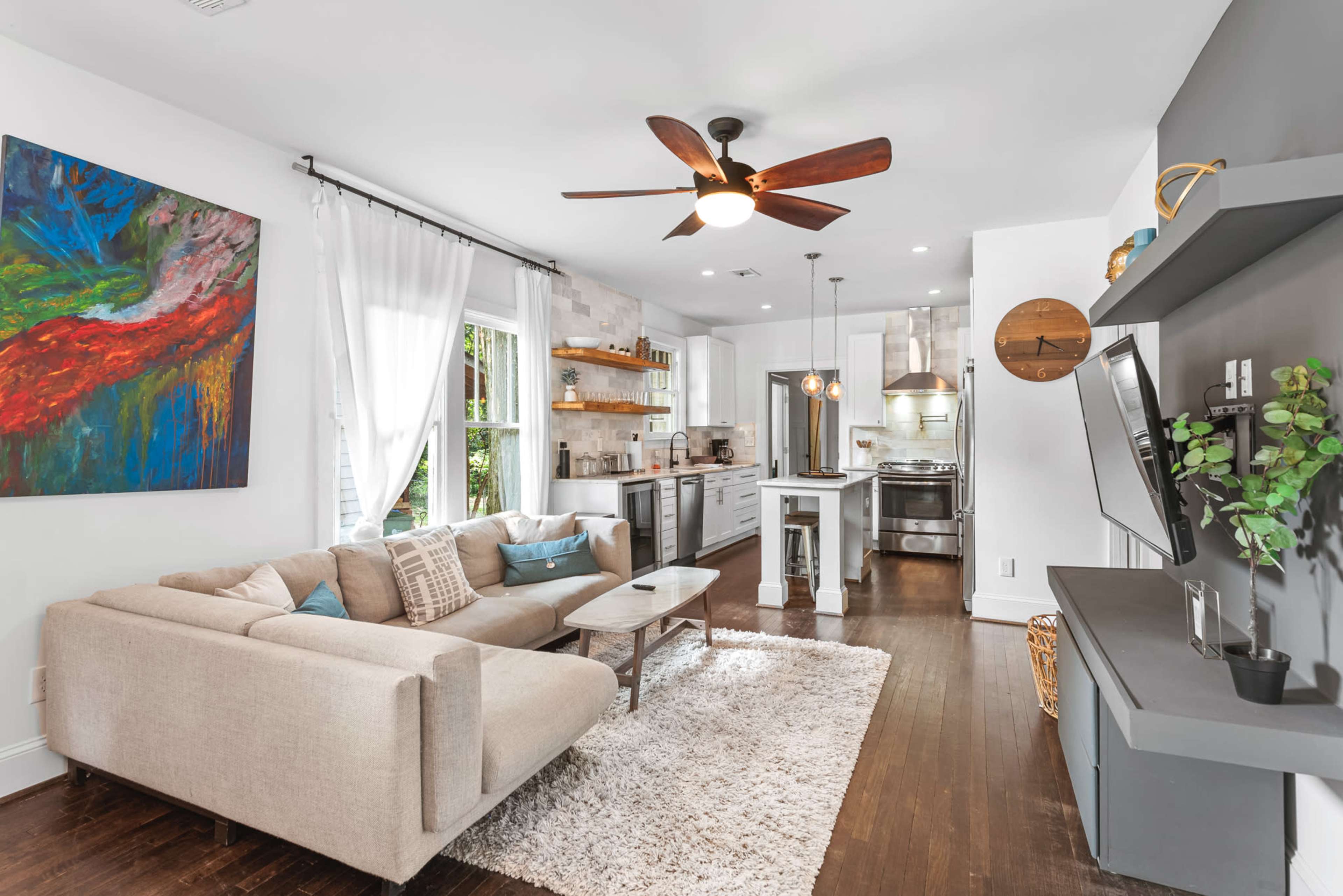 A modern living room with a beige sectional sofa, a wooden coffee table, and a kitchen area featuring stainless steel appliances and open shelving.