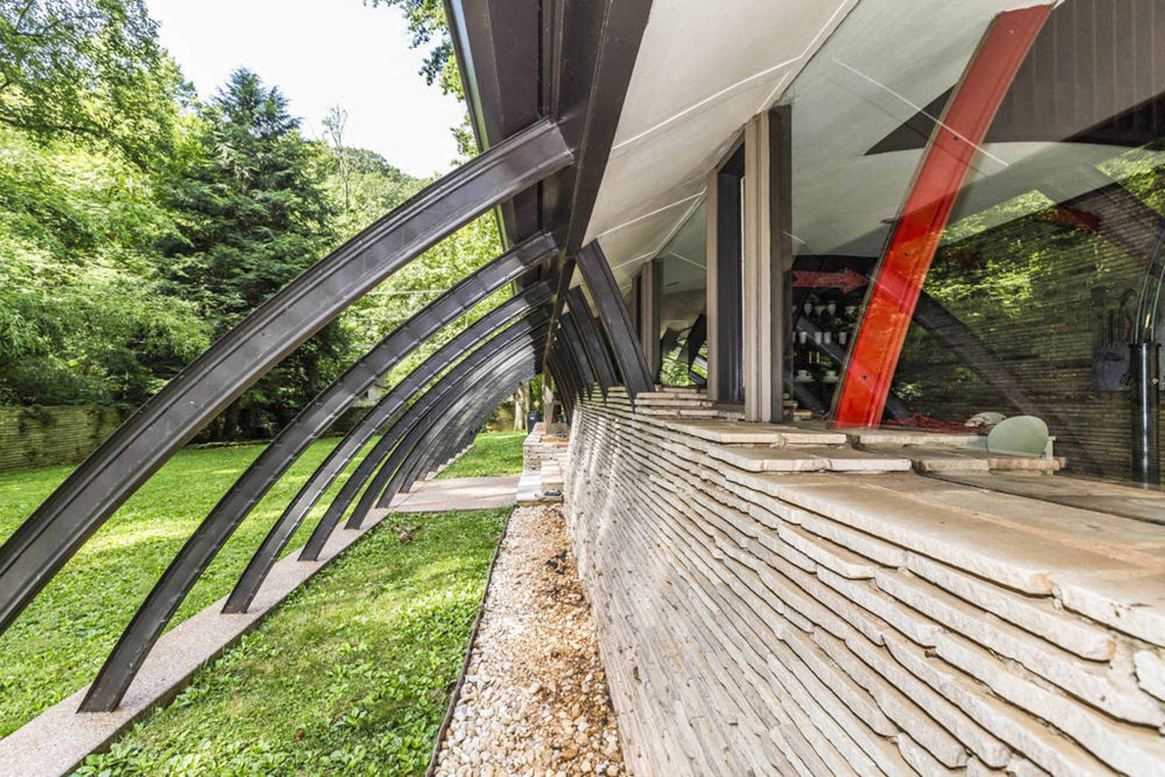 A modern architectural structure with curved metal beams supporting a stone-faced exterior beside a grassy area.
