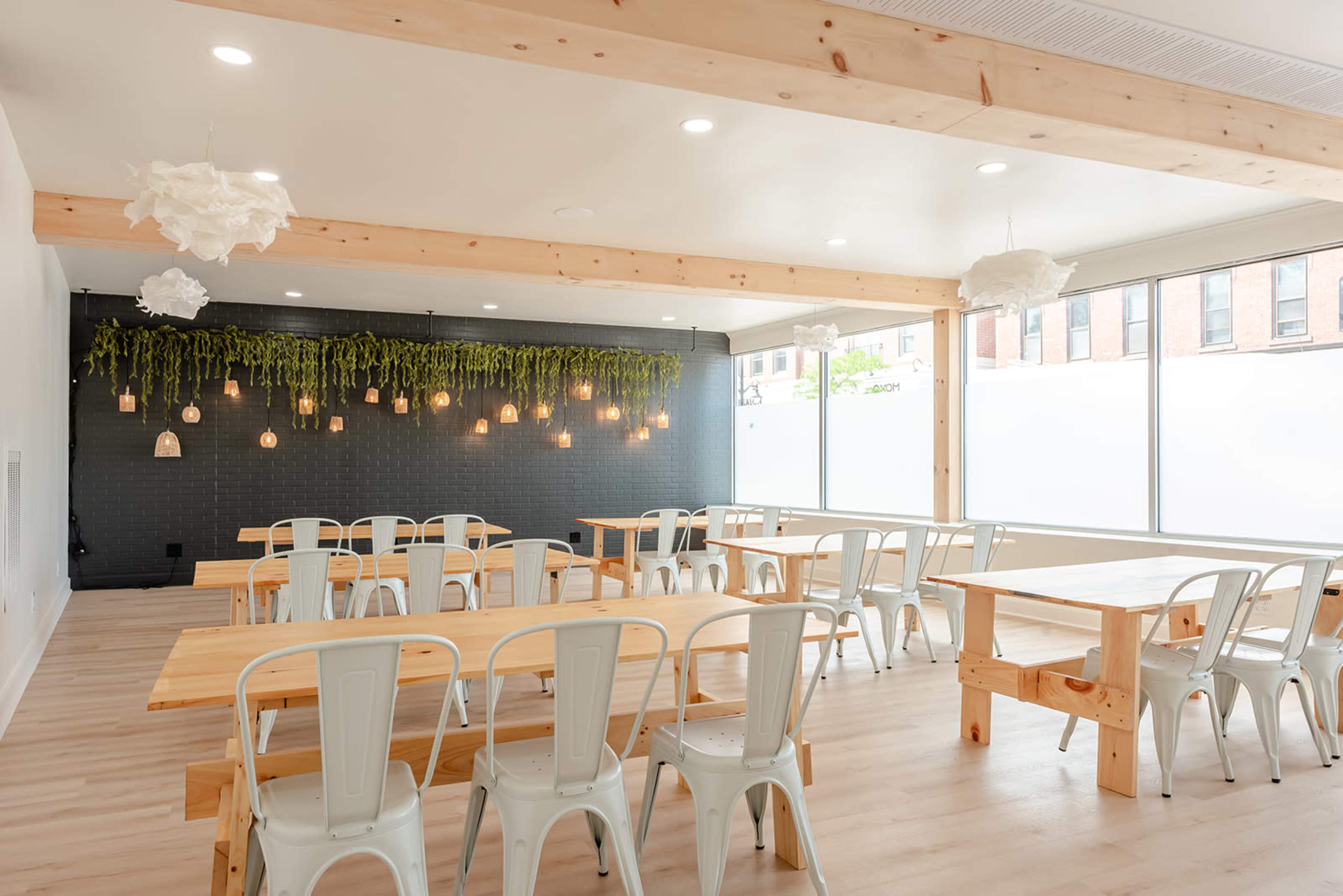 A bright, modern dining area with wooden tables and white metal chairs, featuring hanging lights and greenery along the walls.