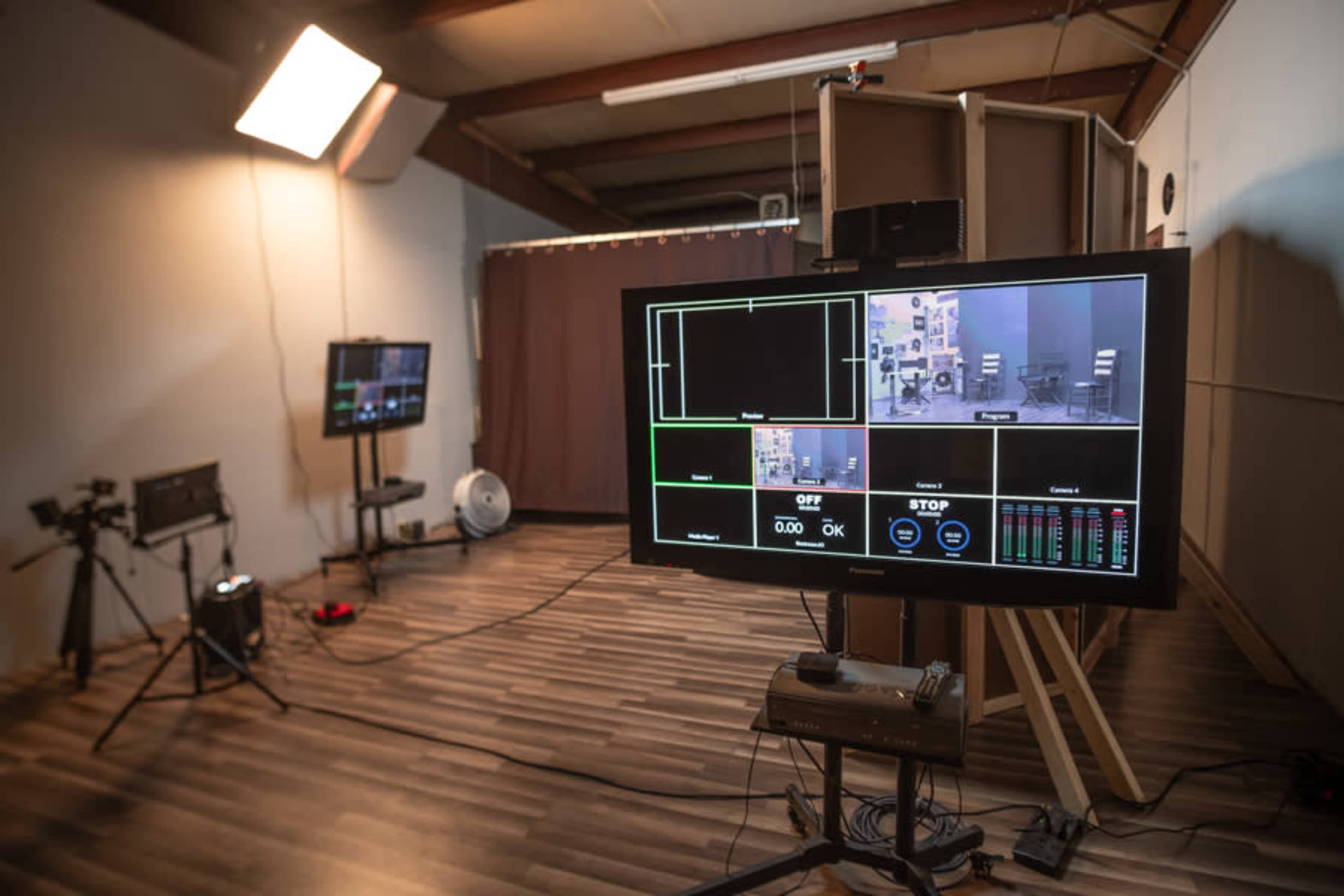 A television studio setup features monitors displaying camera feeds and controls, with lighting equipment and a backdrop in a spacious room.
