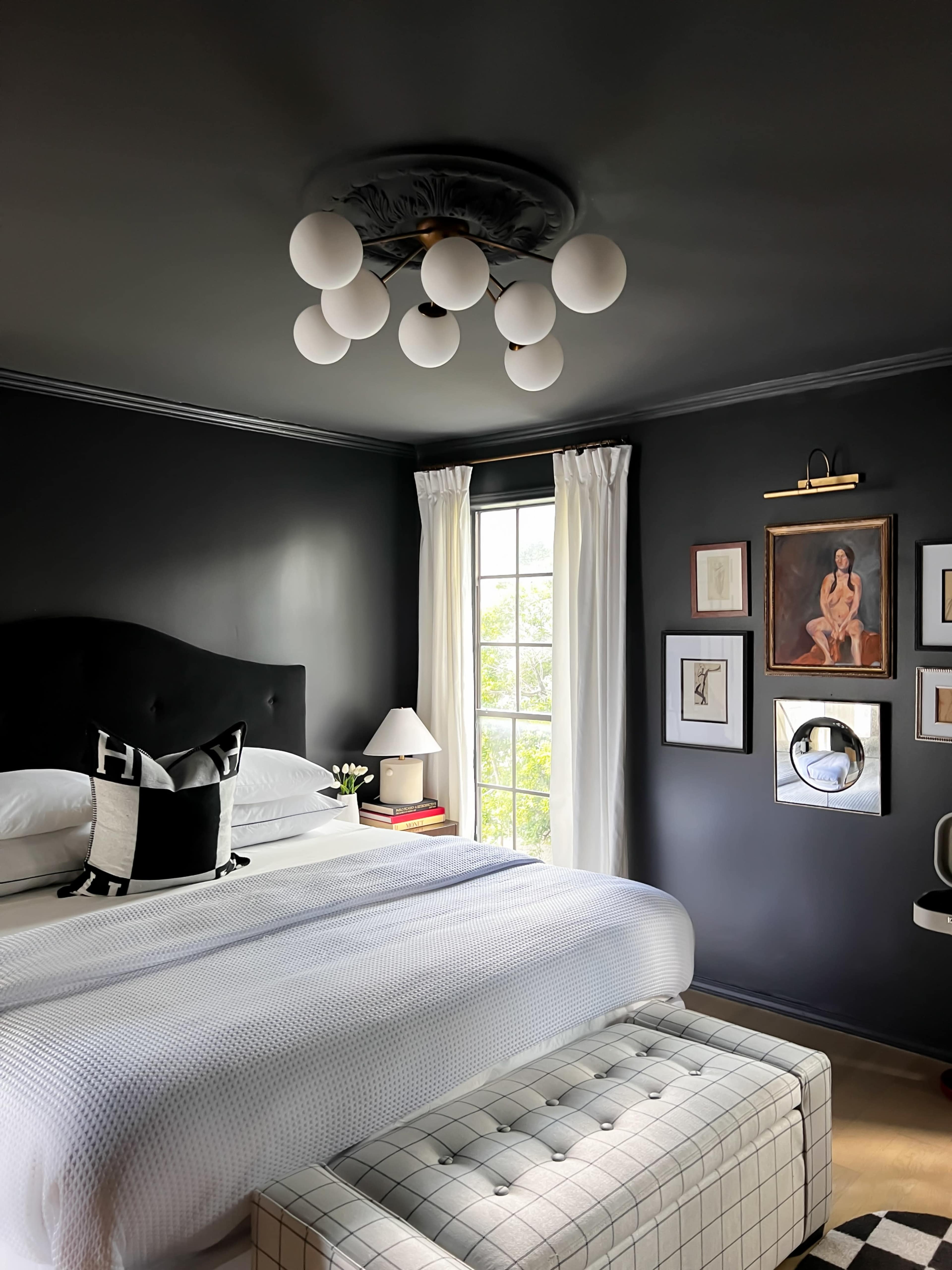 The image shows a modern bedroom featuring a dark ceiling, a large bed with white bedding, a bench at the foot, and various framed artworks on the wall.