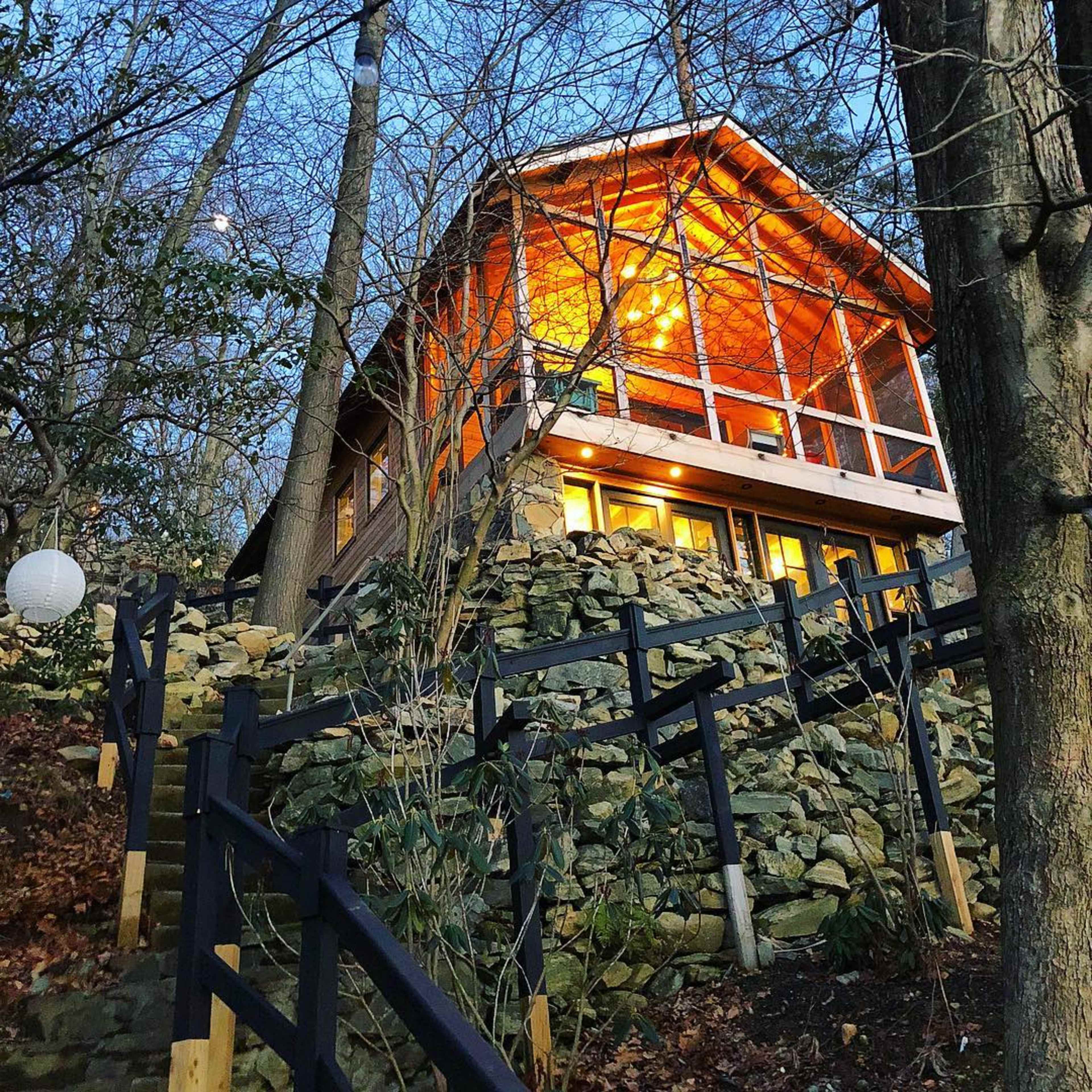 A wooden cabin with bright windows is perched on a rocky hillside surrounded by trees in the evening light.