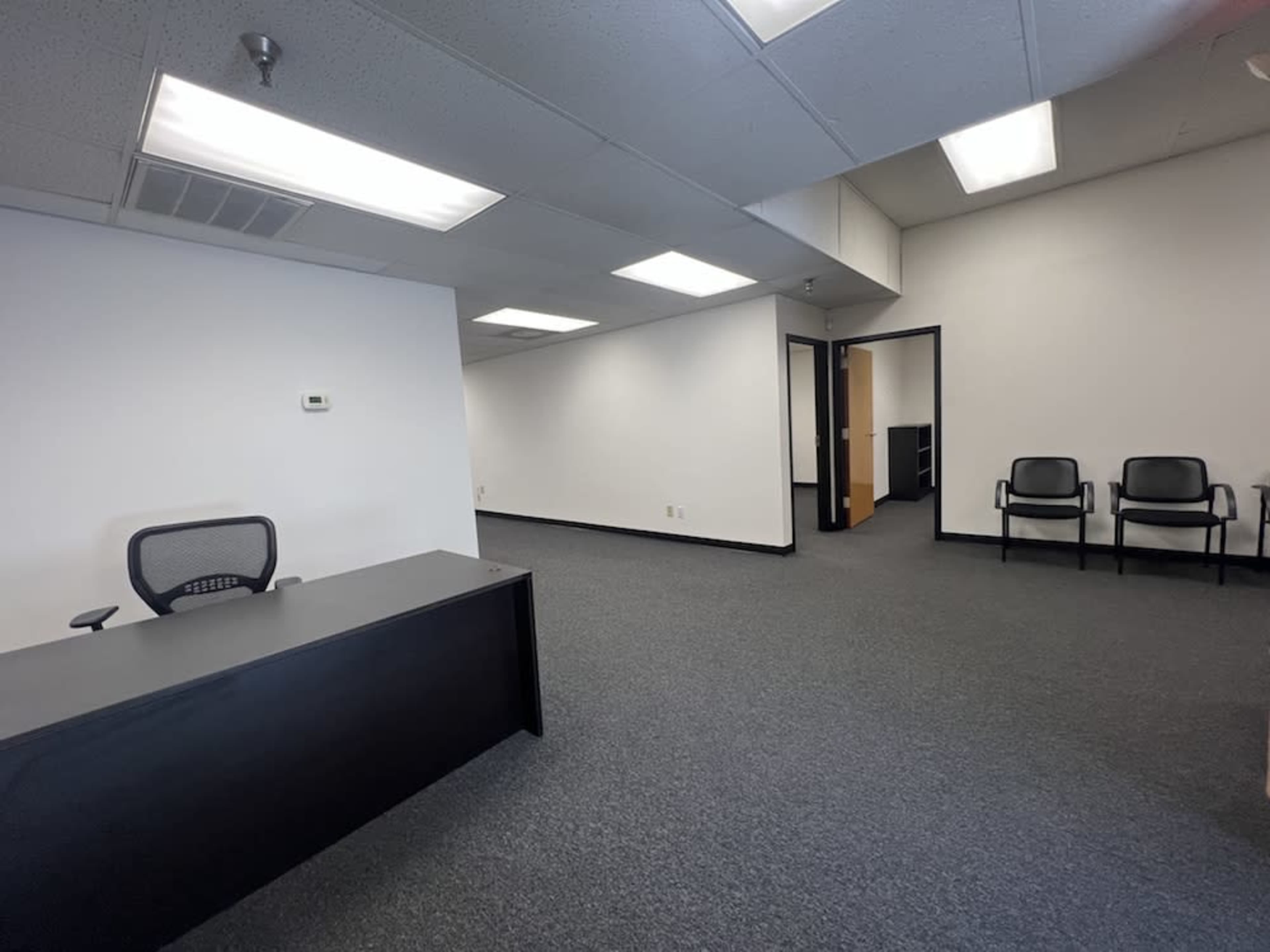 The image shows an empty office space with a black reception desk and a waiting area featuring two chairs, all situated on a carpeted floor under simple fluorescent lighting.