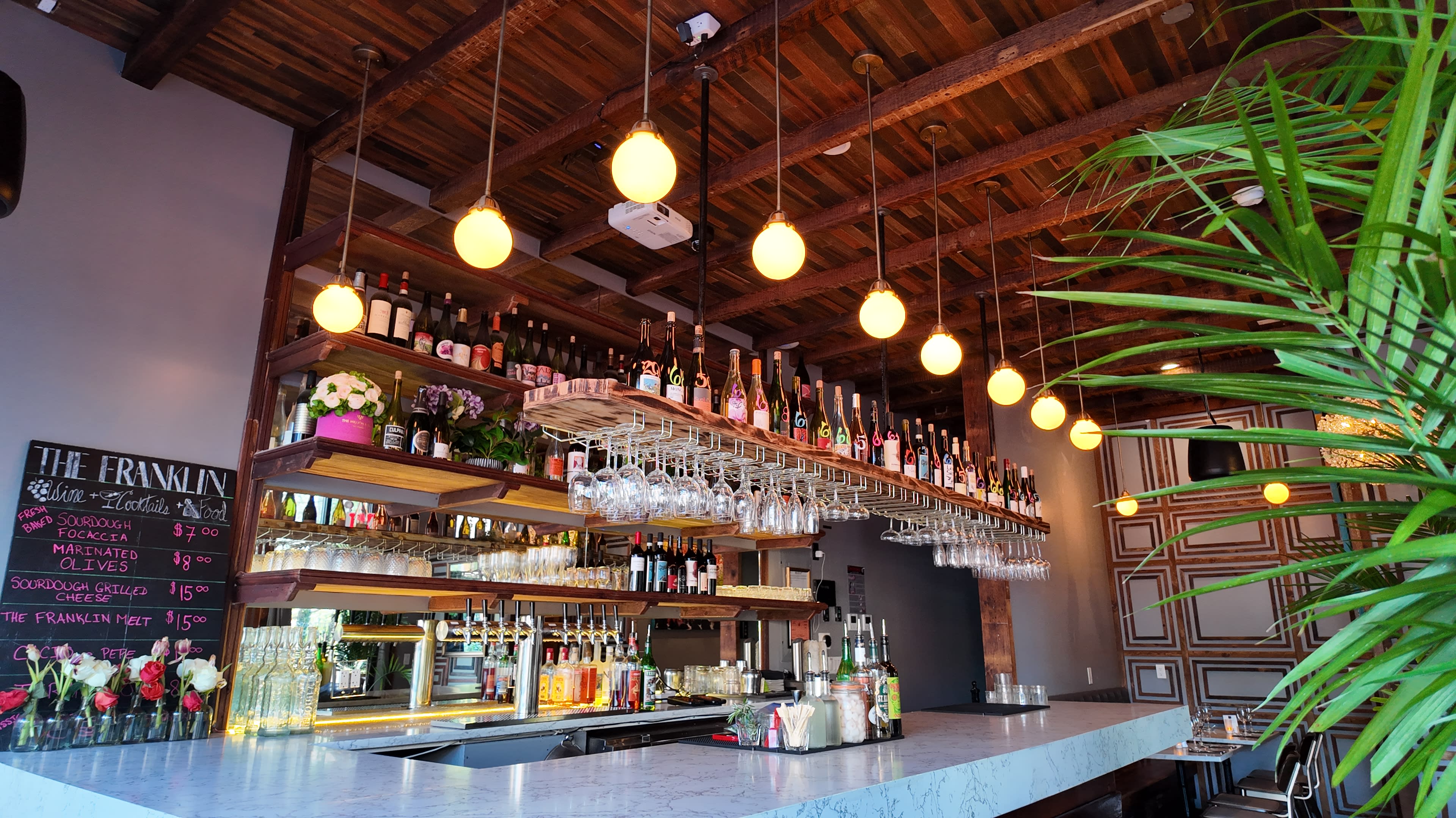 The image shows a modern bar with a wooden ceiling, shelves lined with bottles of wine and liquor, and hanging glassware above a marble countertop.
