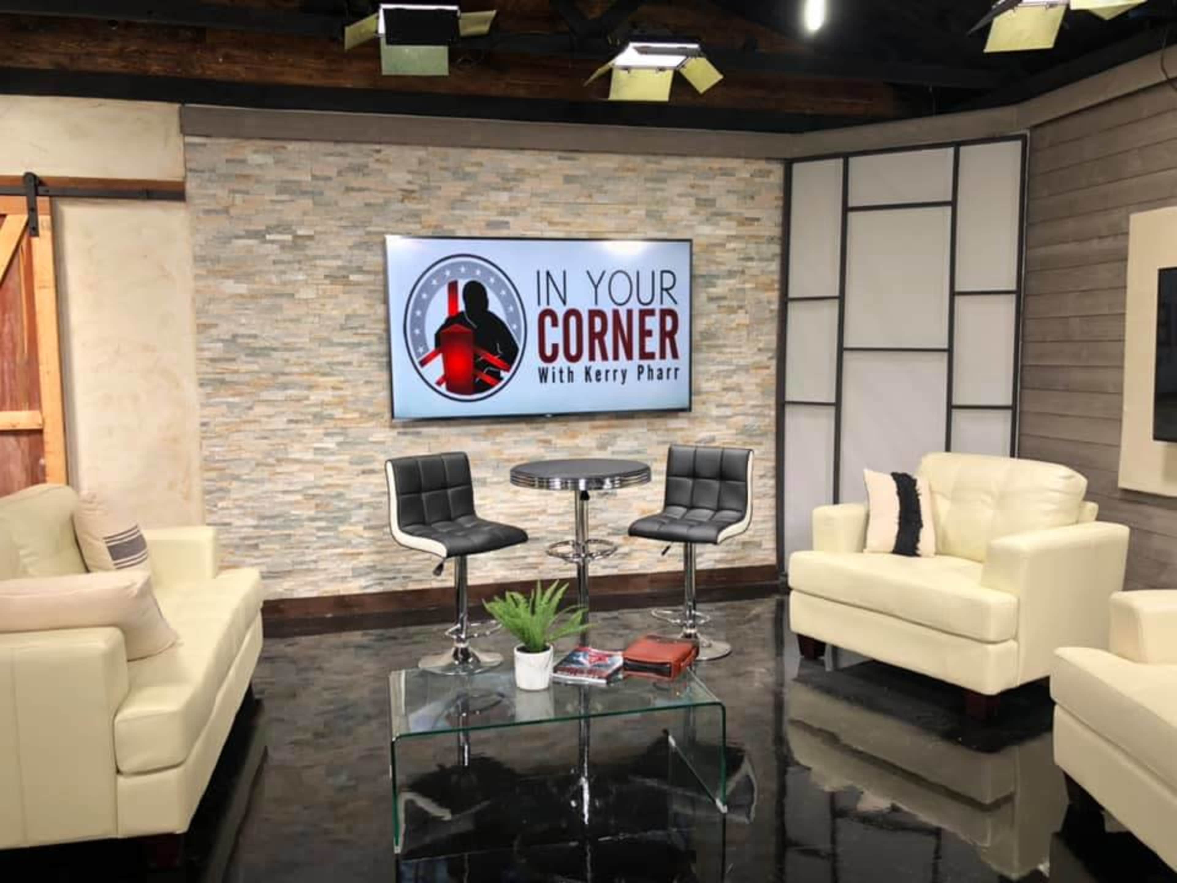 A television studio set with a logo reading "In Your Corner with Kerry Pharr" on a large screen, featuring two black chairs around a round table and light-colored sofas.