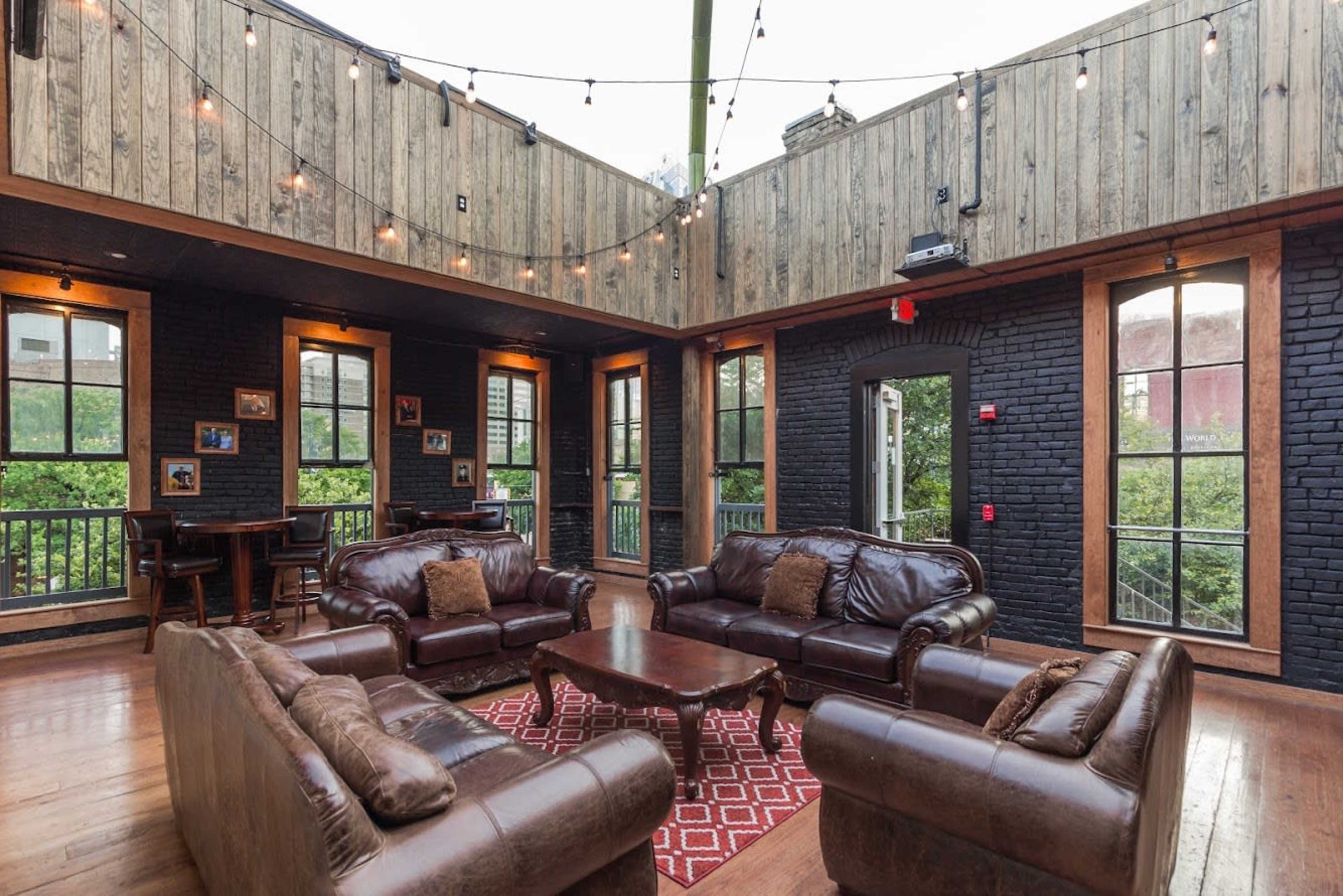 A cozy lounge area with leather sofas arranged around a central coffee table, illuminated by string lights and featuring large windows that provide views of greenery outside.