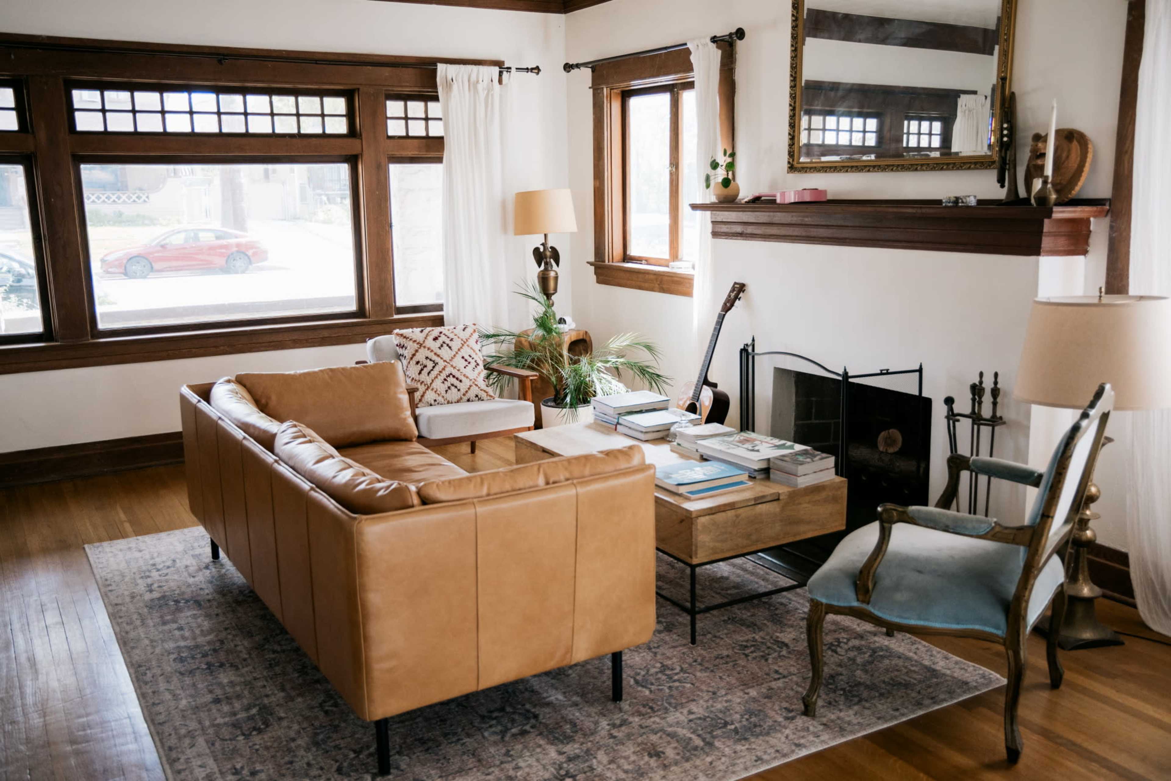 The living room features a tan leather sofa, a wooden coffee table stacked with books, and large windows allowing natural light to fill the space.