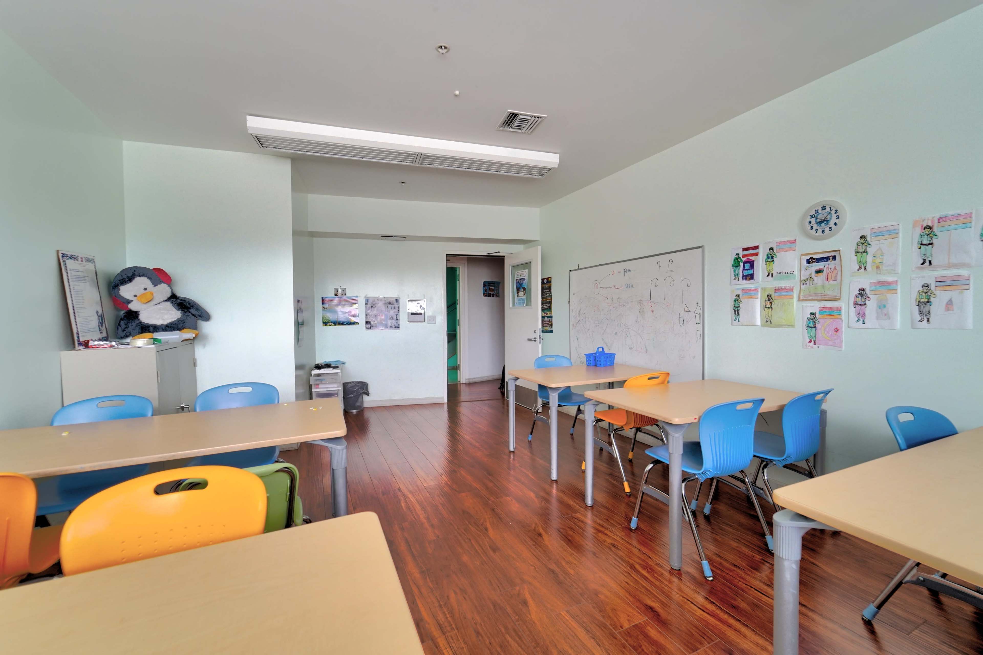 A classroom with wooden floors, light green walls, several tables with colorful chairs, and a whiteboard covered in drawings and posters.