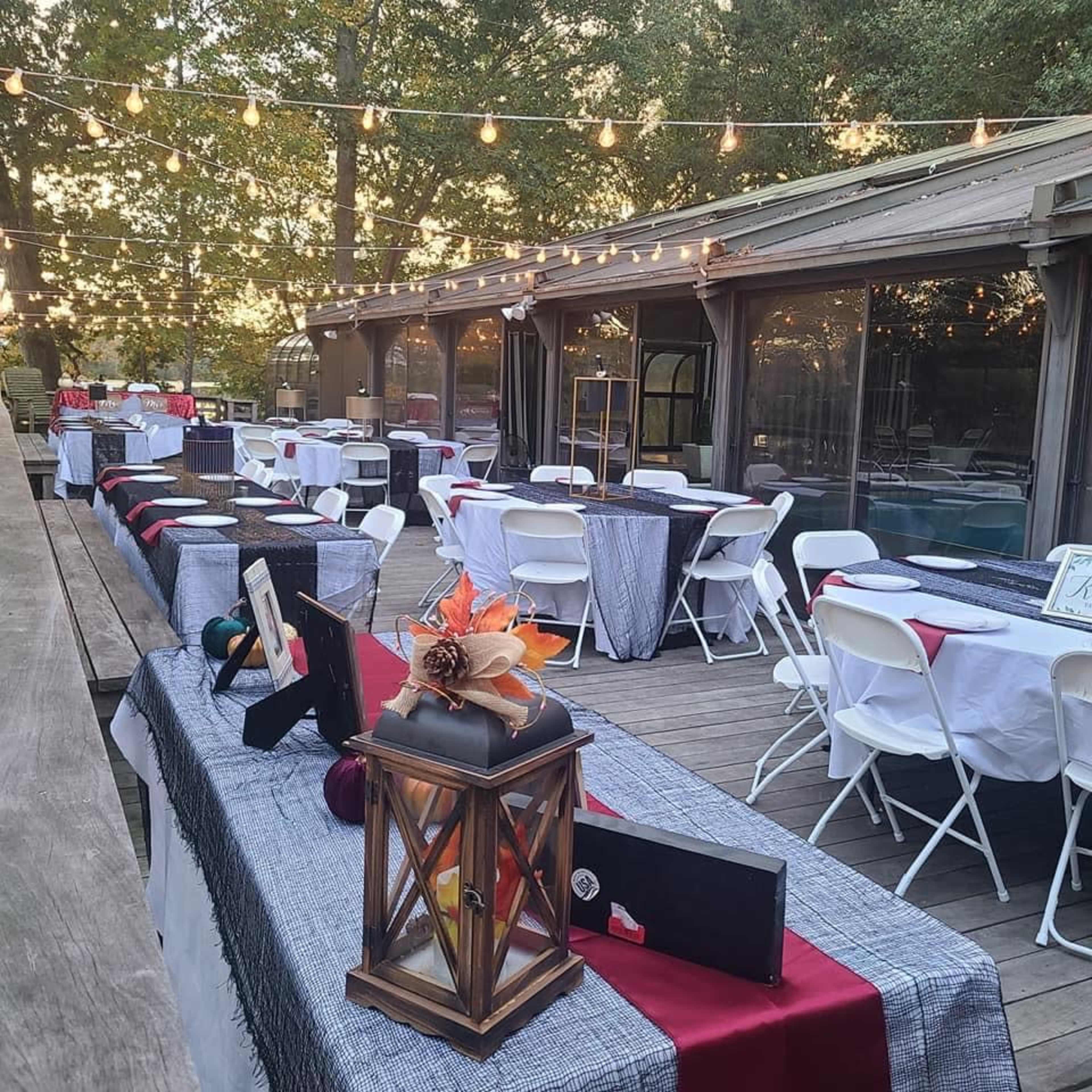 A series of tables are arranged outside, adorned with tablecloths and decorations, next to a building with large windows and hanging lights overhead.