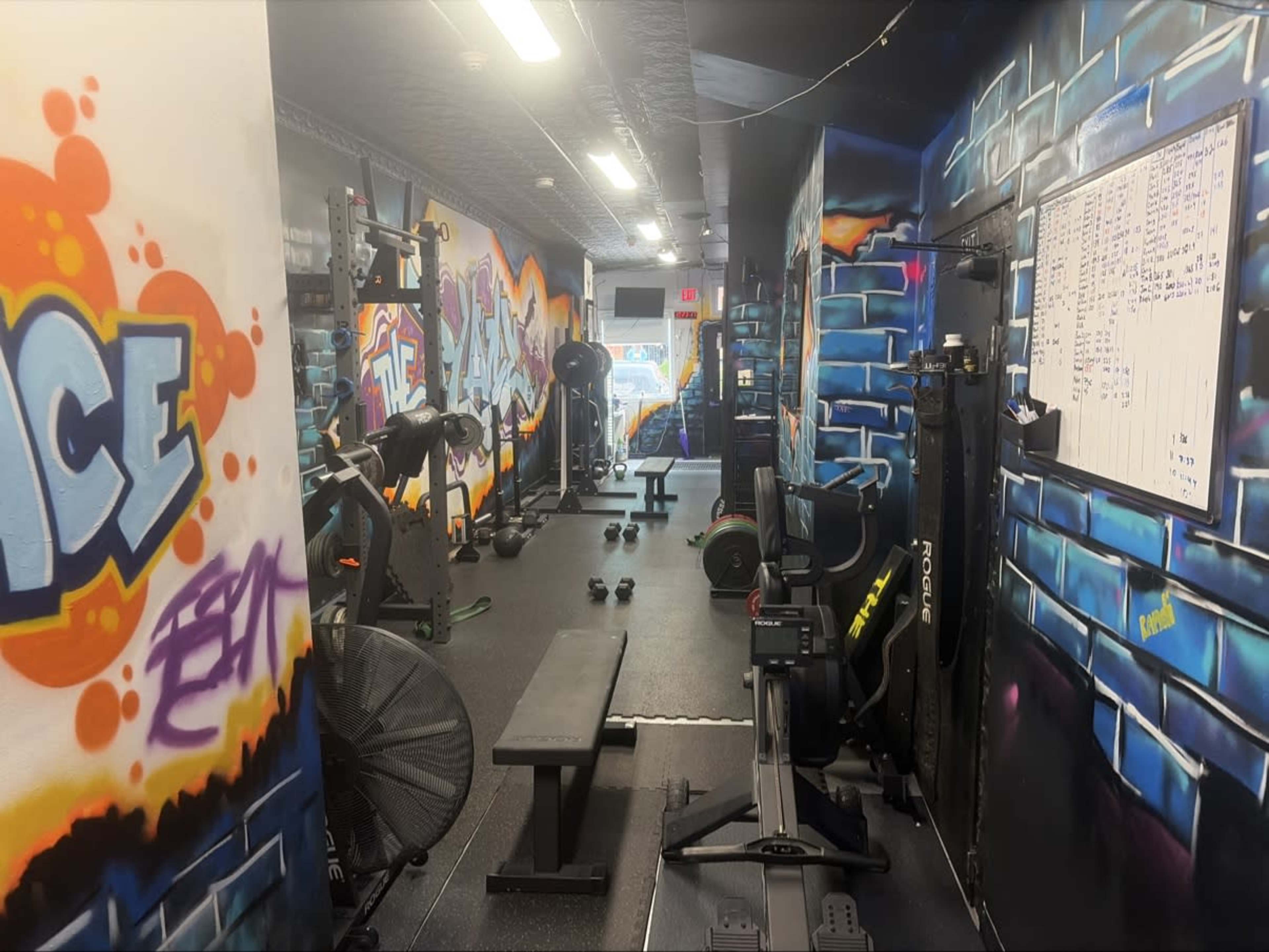 The image shows a gym interior featuring workout equipment, vibrant graffiti art on the walls, and a whiteboard displaying workout schedules.