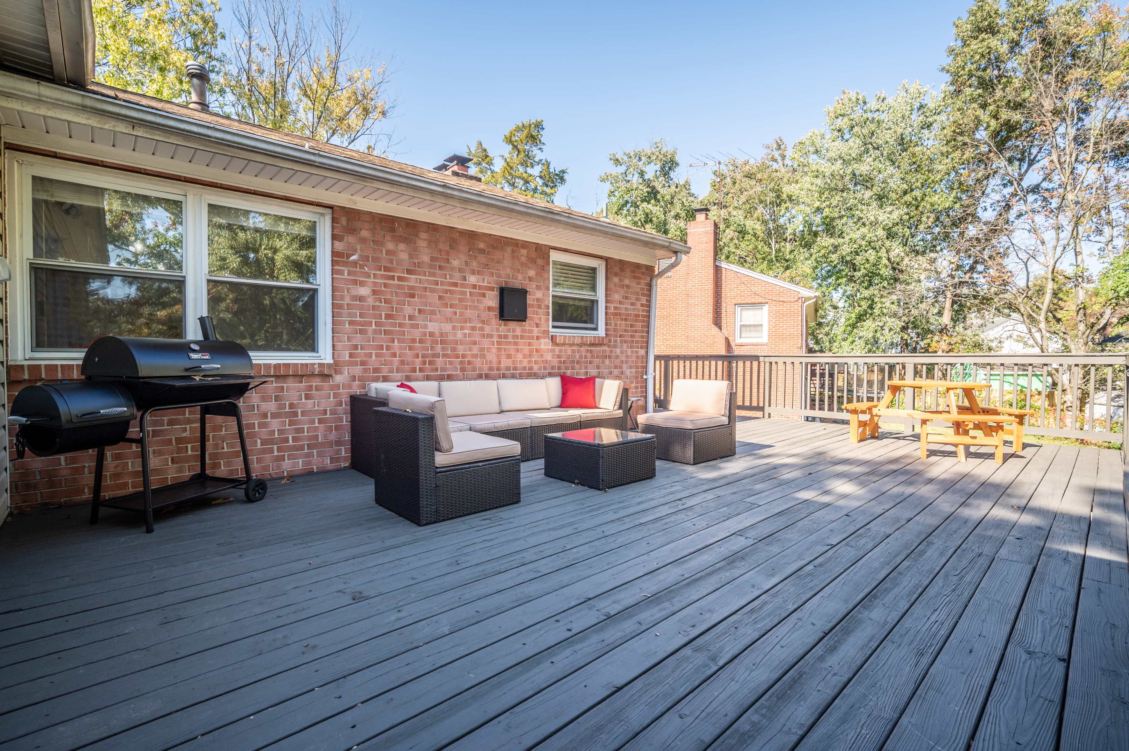 A wooden deck features a seating area with a sectional sofa and coffee table, alongside a barbecue grill and a dining table.