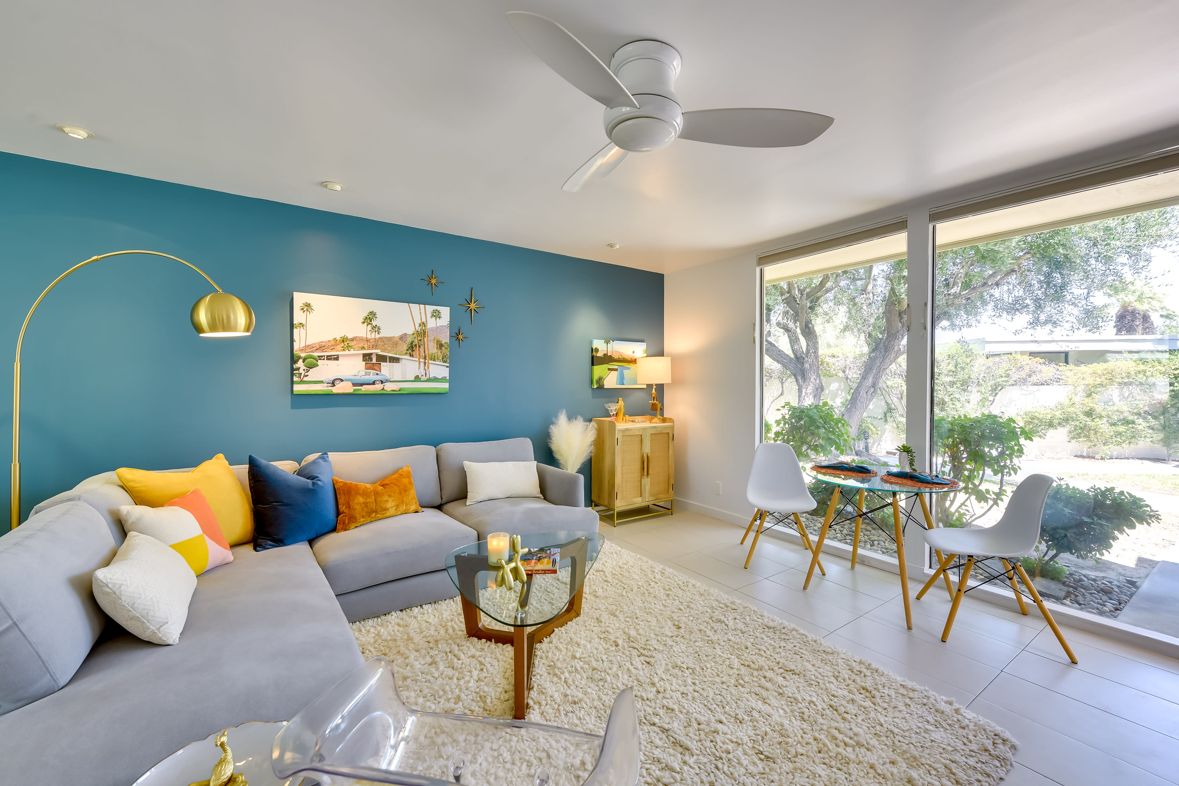 The image shows a modern living room with a sectional couch, colorful decorative pillows, a coffee table, and a dining area featuring a small table and chairs by large windows.