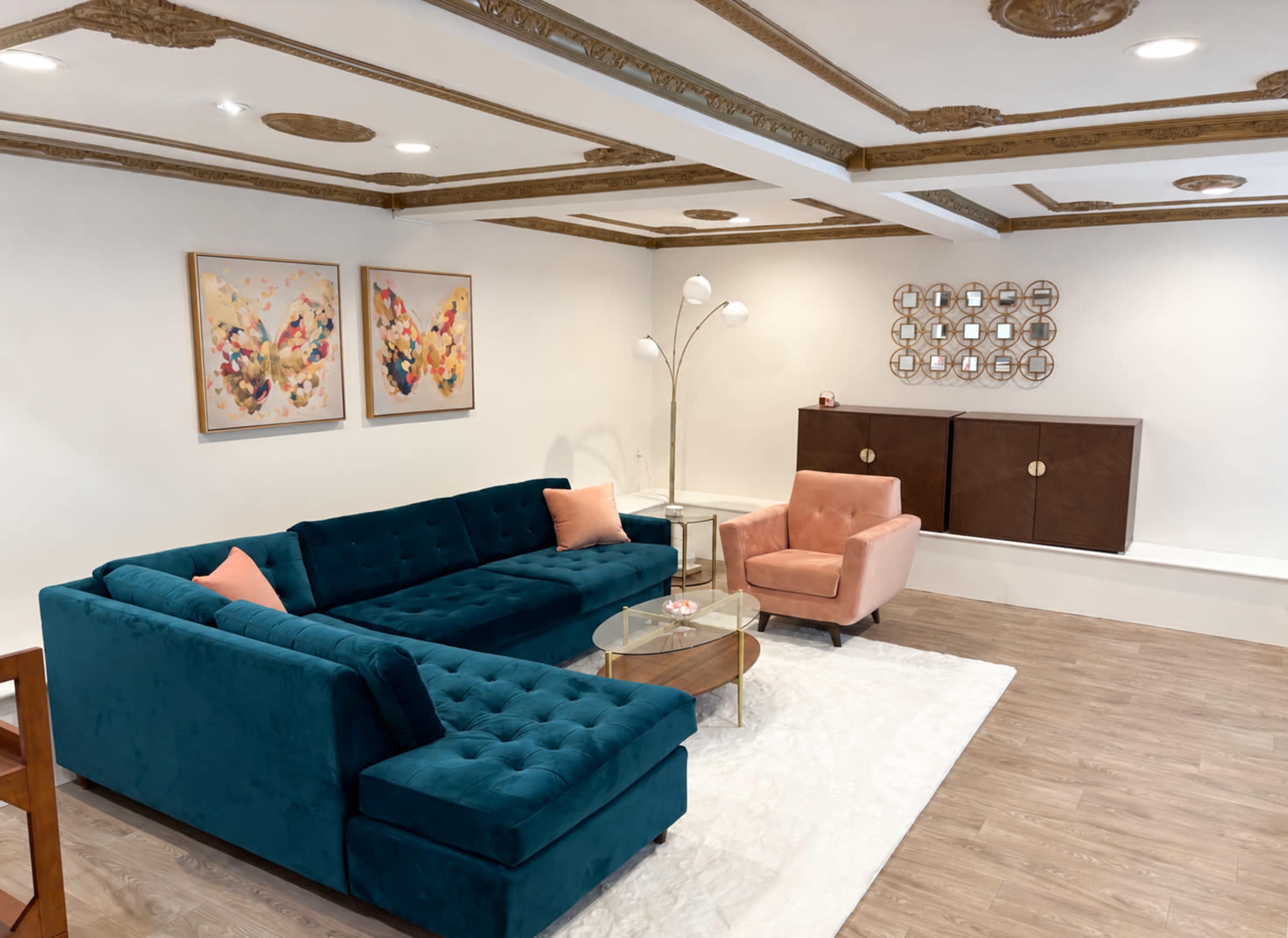 The image displays a modern living room featuring a teal sectional sofa, a pink accent chair, a glass coffee table, and two framed artworks on the wall.