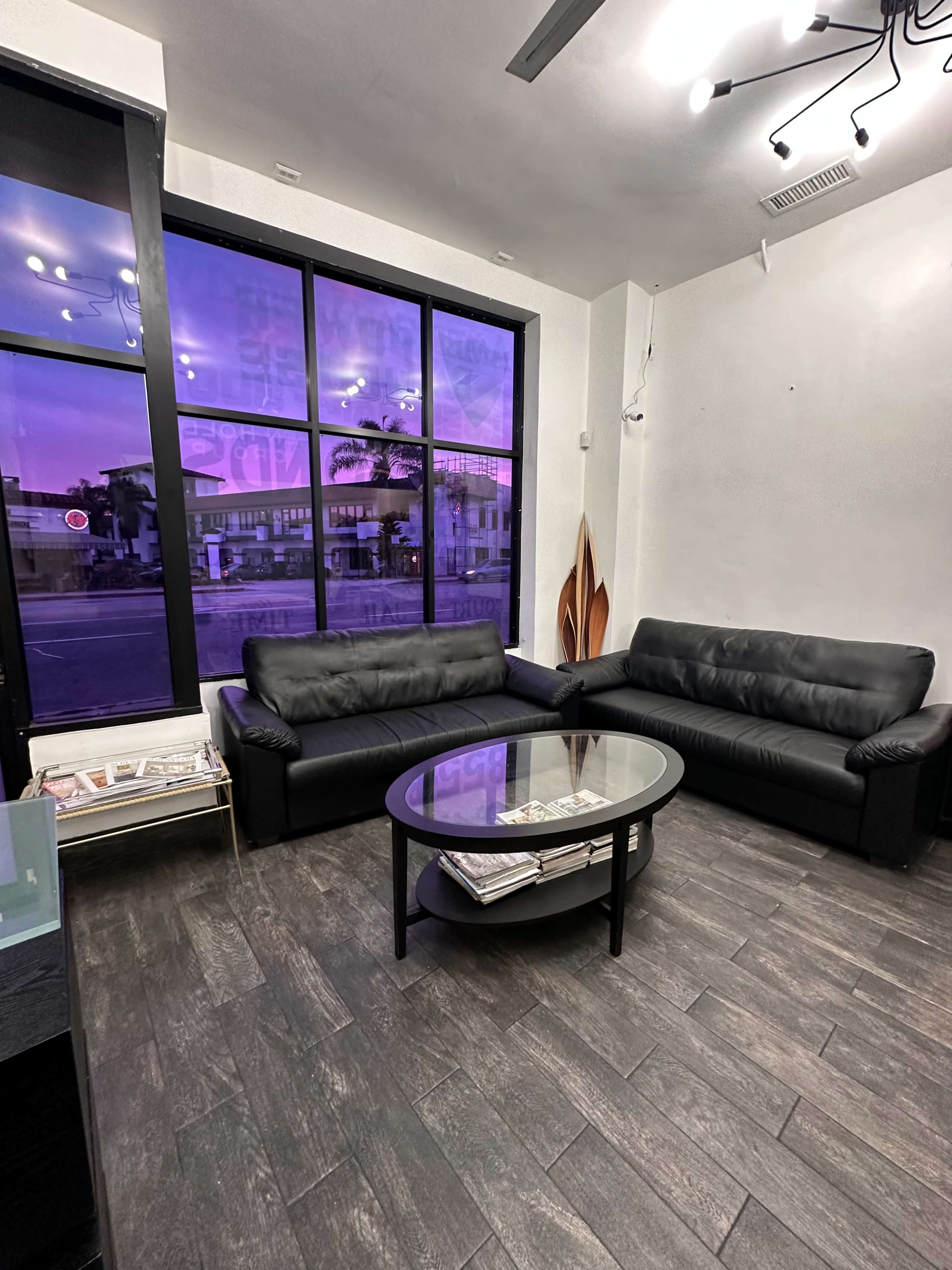 The image shows a modern waiting area with two black leather sofas and a glass coffee table, illuminated by purple light coming through large windows.
