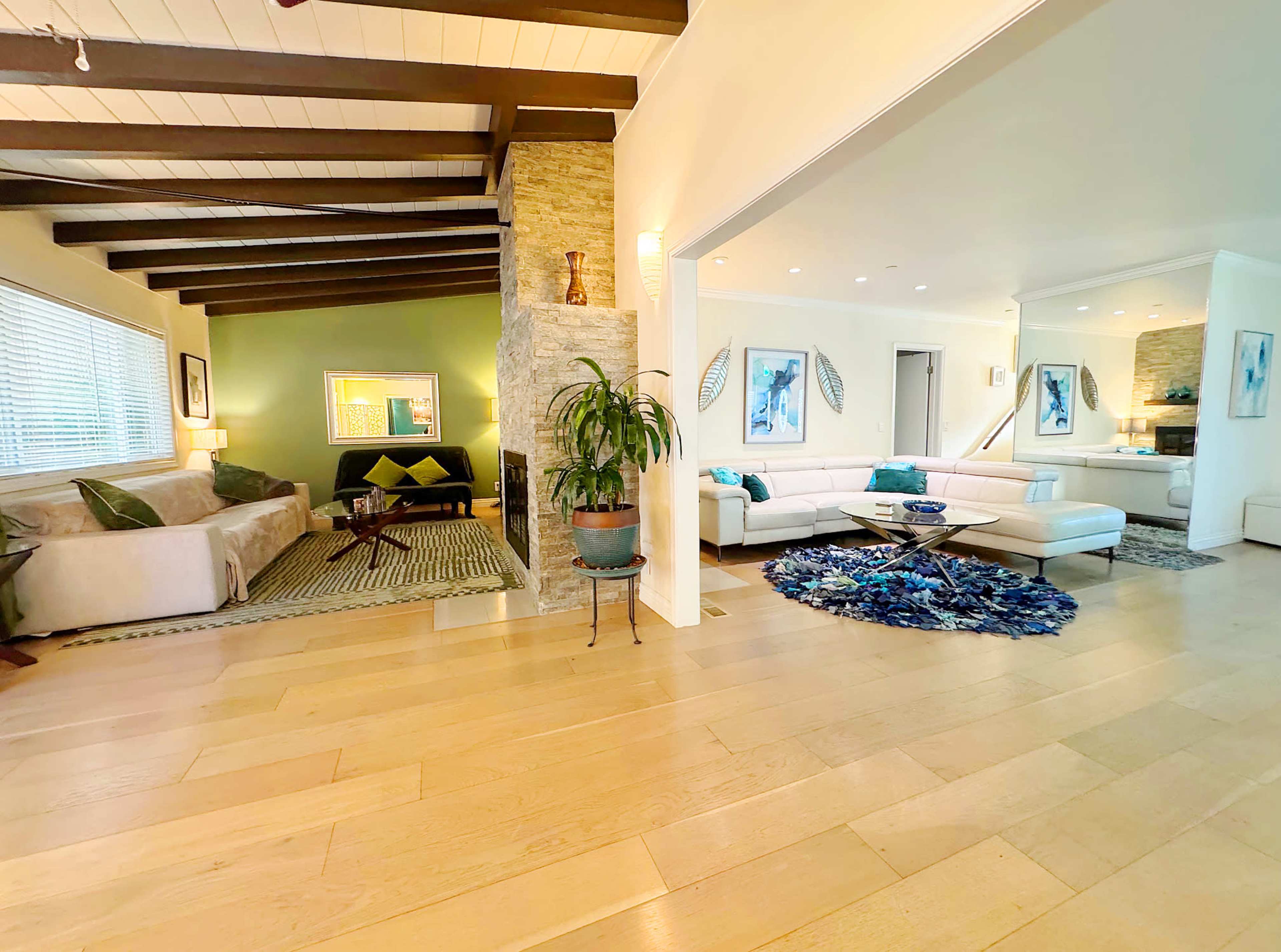 The image shows a spacious living room with a mix of modern and cozy seating arrangements, a stone fireplace, and decorative elements, including plants and art pieces.