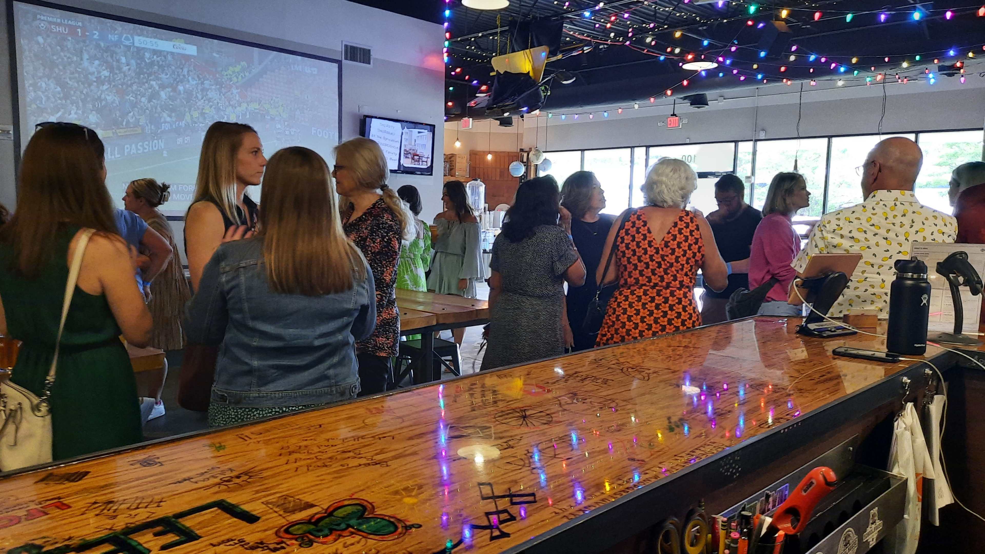 A group of people is socializing in a brightly lit bar with colorful string lights above them.