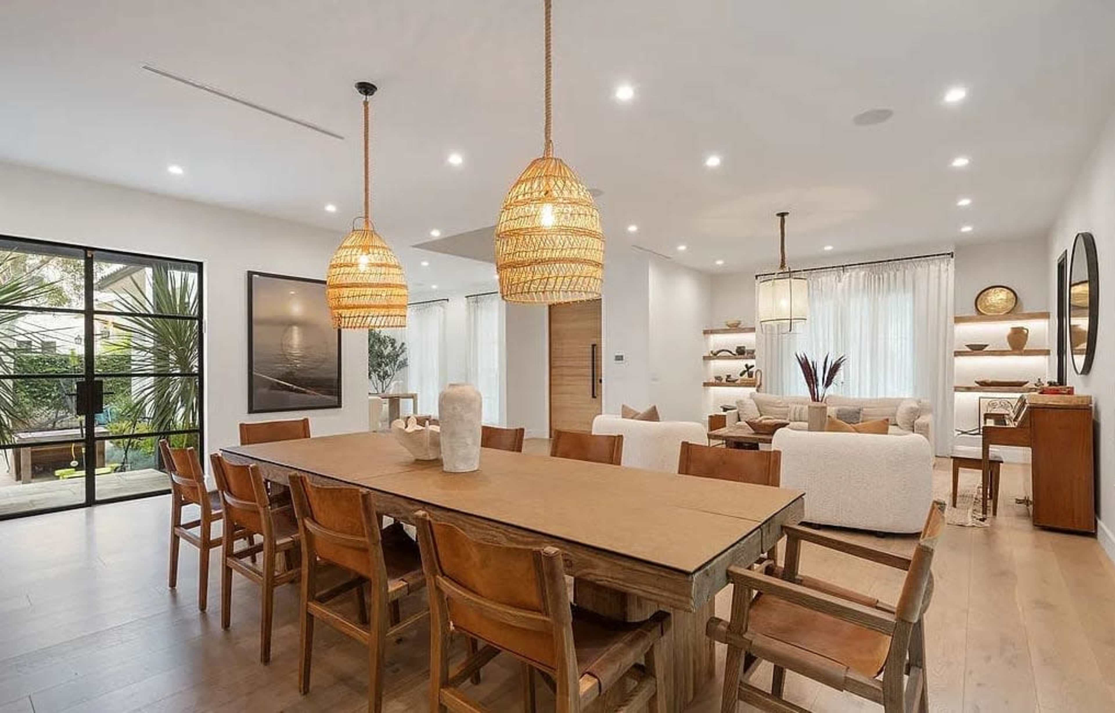 The image shows a modern dining area with a long wooden table surrounded by chairs, pendant lights hanging above, and a spacious open layout that connects to a living space.