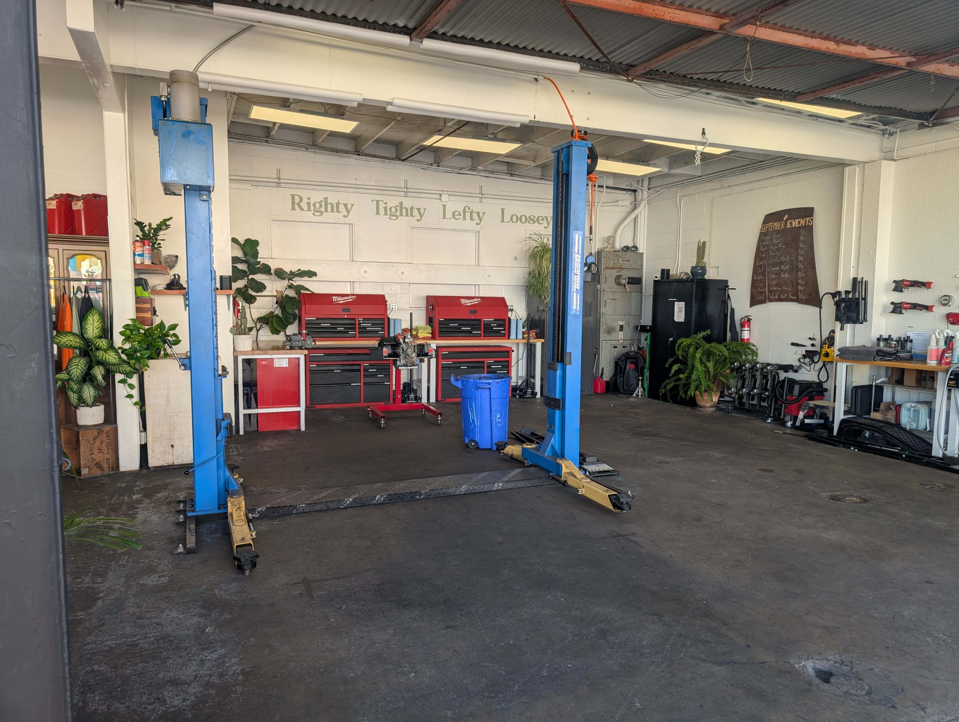 The image shows an automotive workshop with a blue car lift in the center, red toolboxes along the wall, and various equipment and plants scattered around.