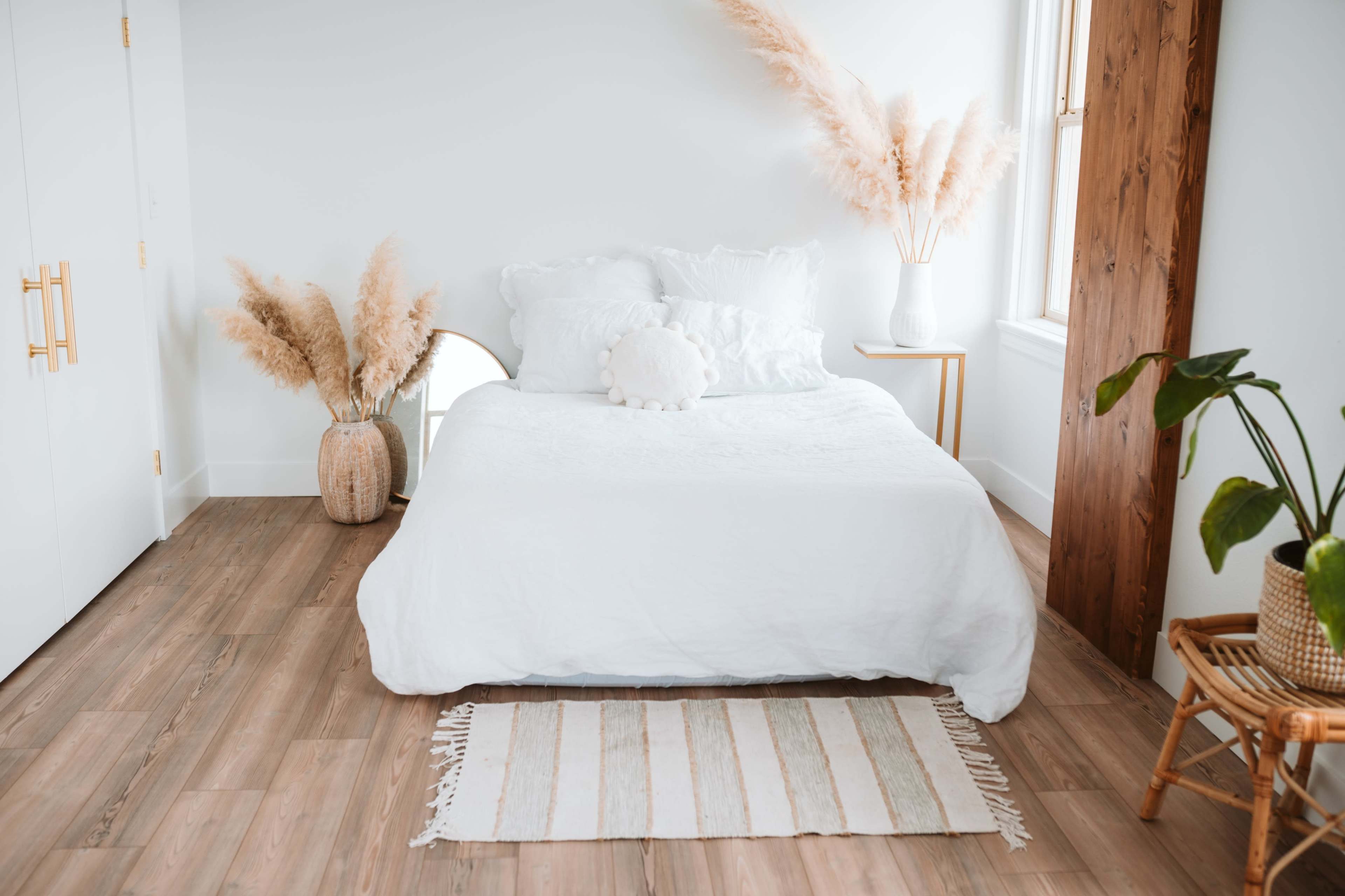 A minimalistic bedroom features a white bed with decorative pillows, a small side table, and plants placed near large windows.