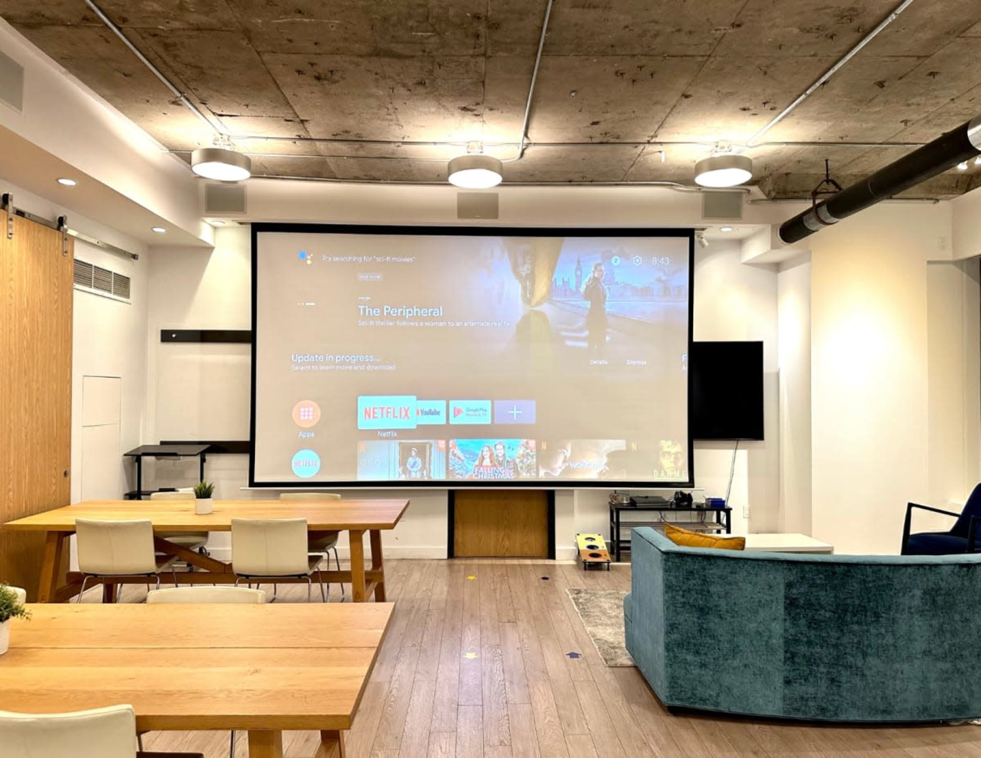 A large screen displaying a streaming service interface is mounted on a wall in a modern room with wooden tables, a blue couch, and exposed ceilings.
