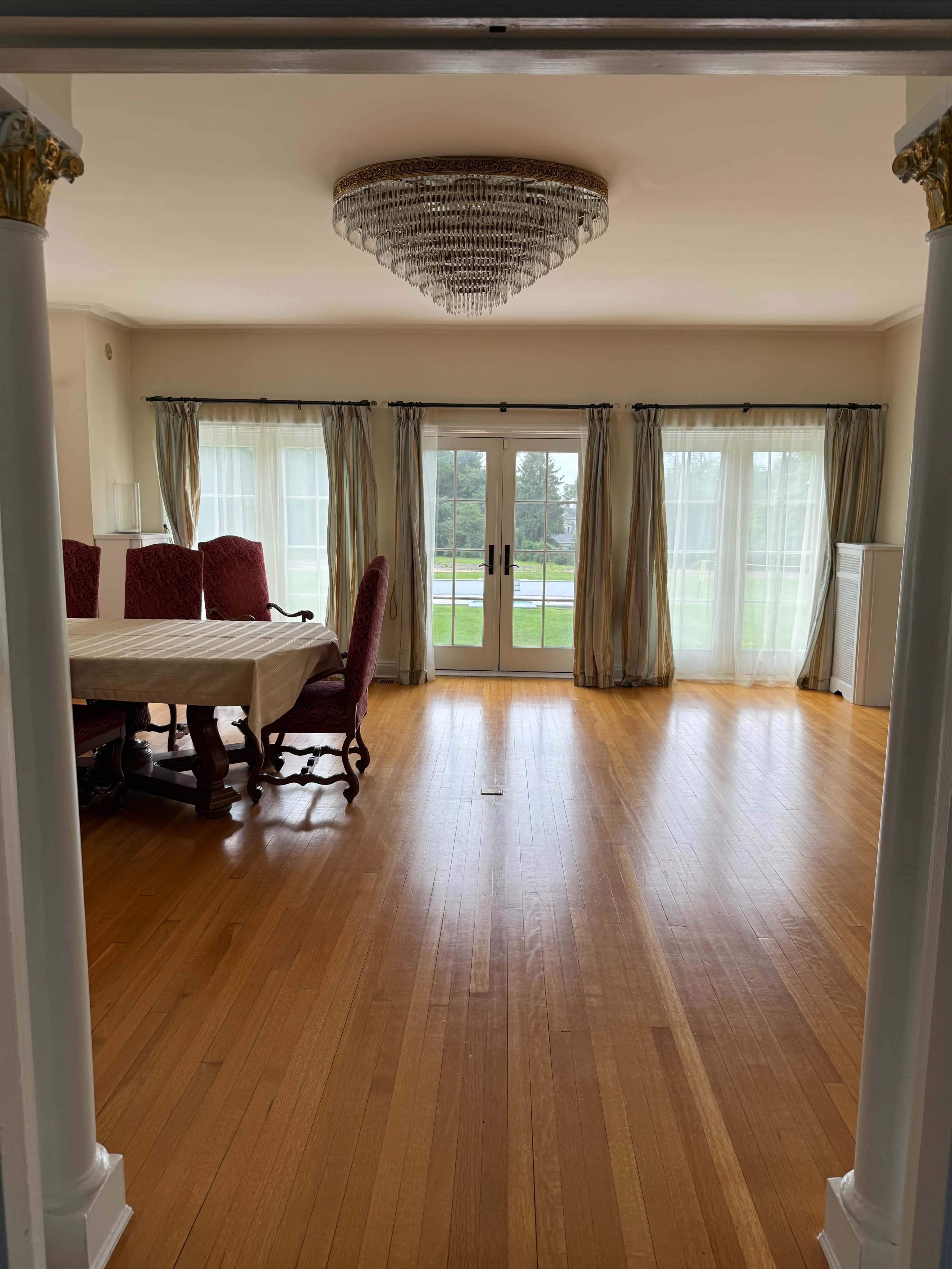 The image shows a spacious room with wood flooring, featuring a dining table and chairs, large windows with sheer curtains, and a chandelier hanging from the ceiling.