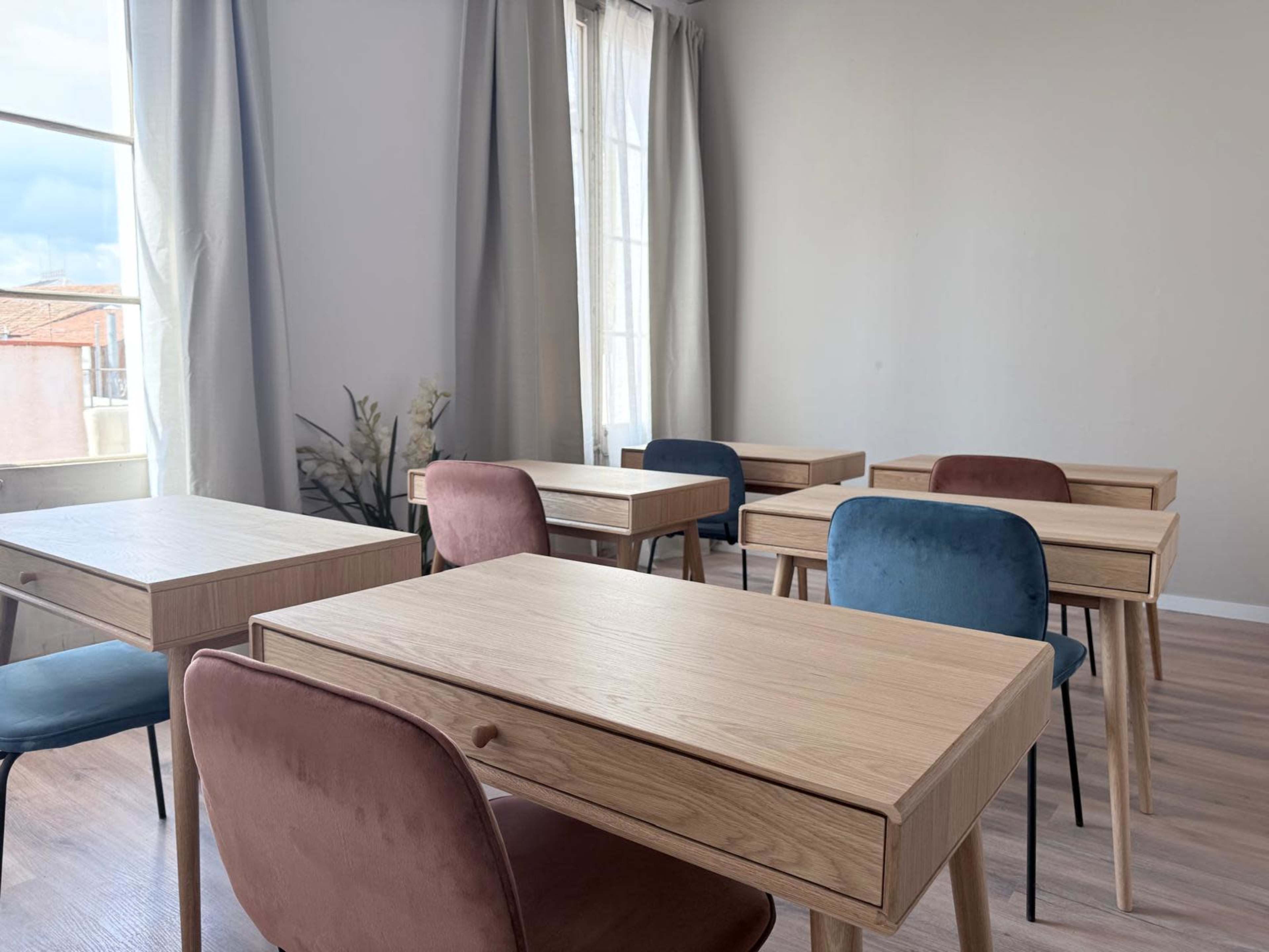 The image shows a modern room with several wooden tables and chairs arranged around a light-colored space with large windows.