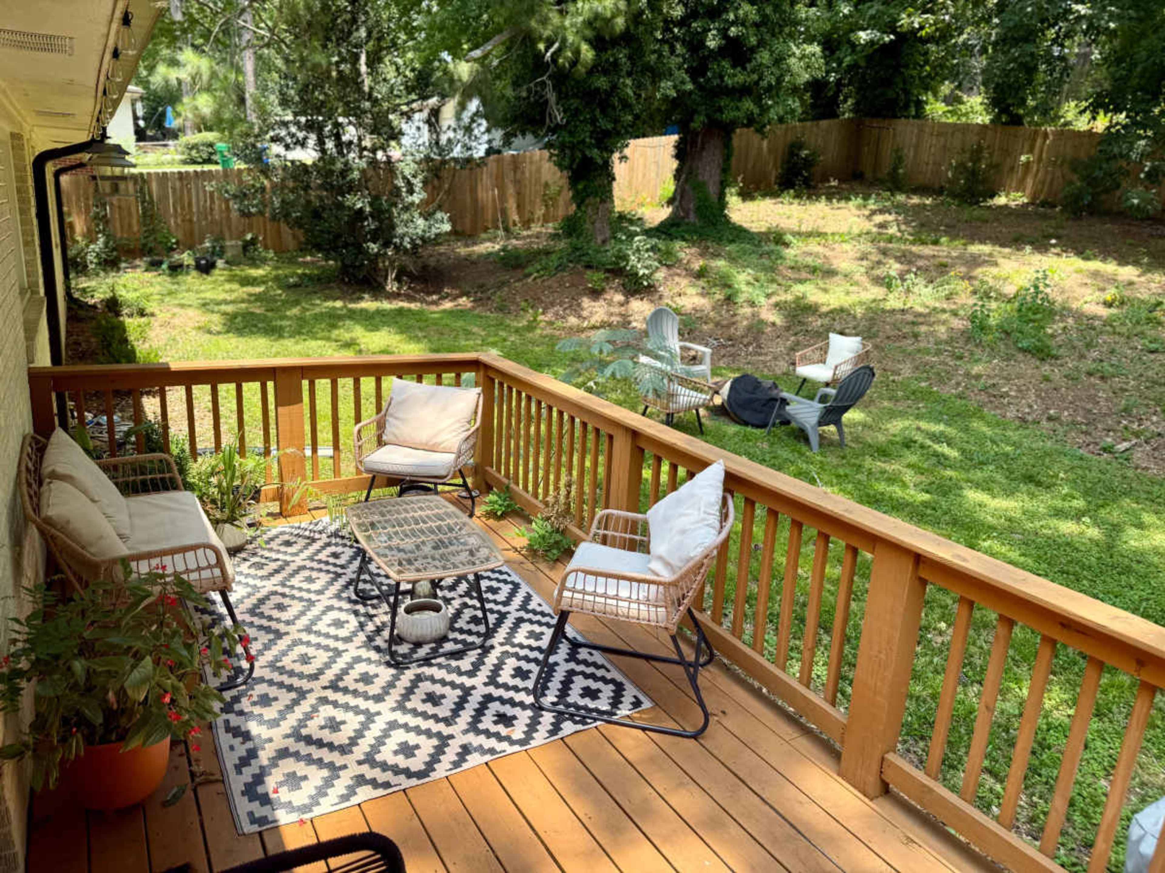 A wooden deck is furnished with a seating area and a patterned rug, overlooking a grassy backyard surrounded by trees.