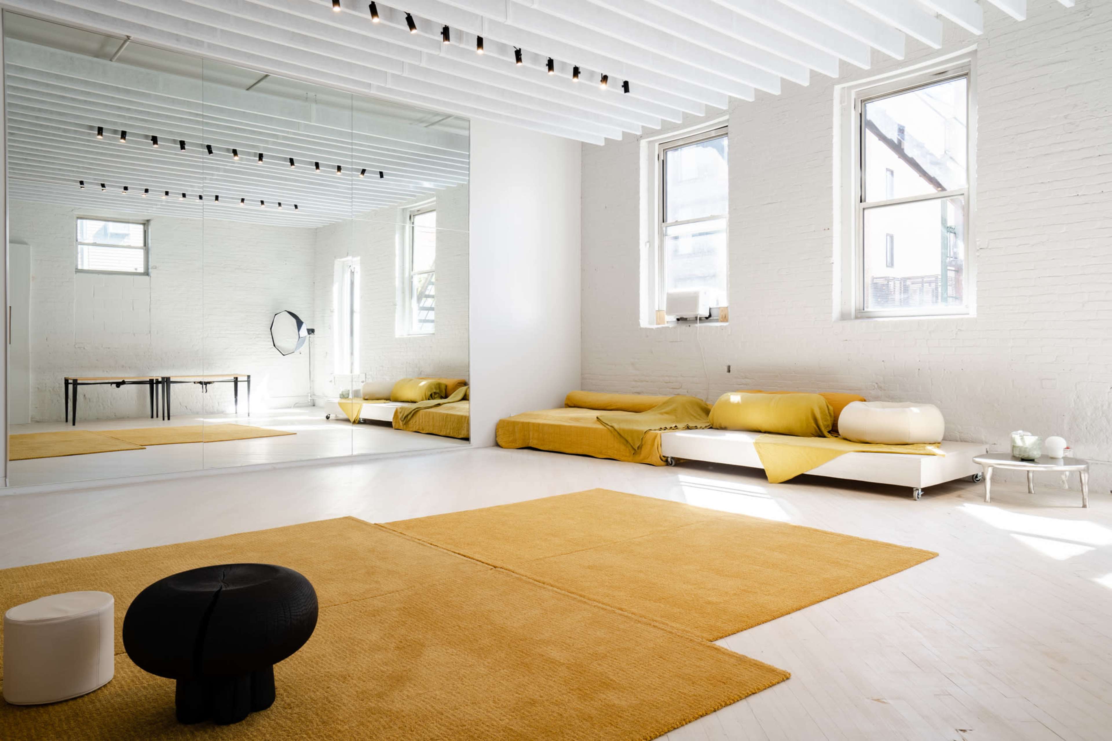 The image shows a minimalist interior space with a large mirror, a white sofa covered in yellow throws, and a beige rug on the floor.