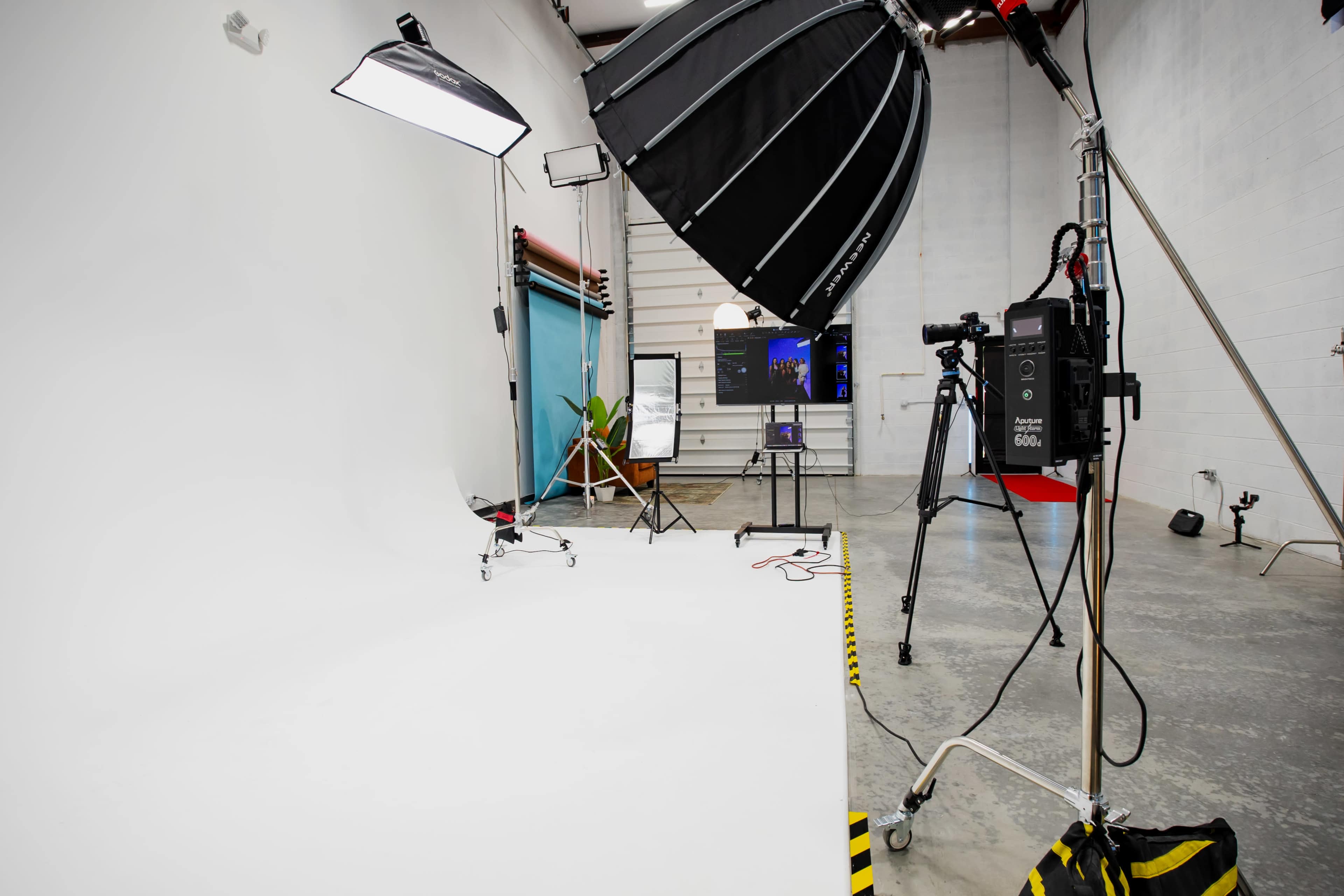 The image shows a photography studio setup featuring softbox lights, a backdrop, a camera on a tripod, and a table with a laptop and a plant.