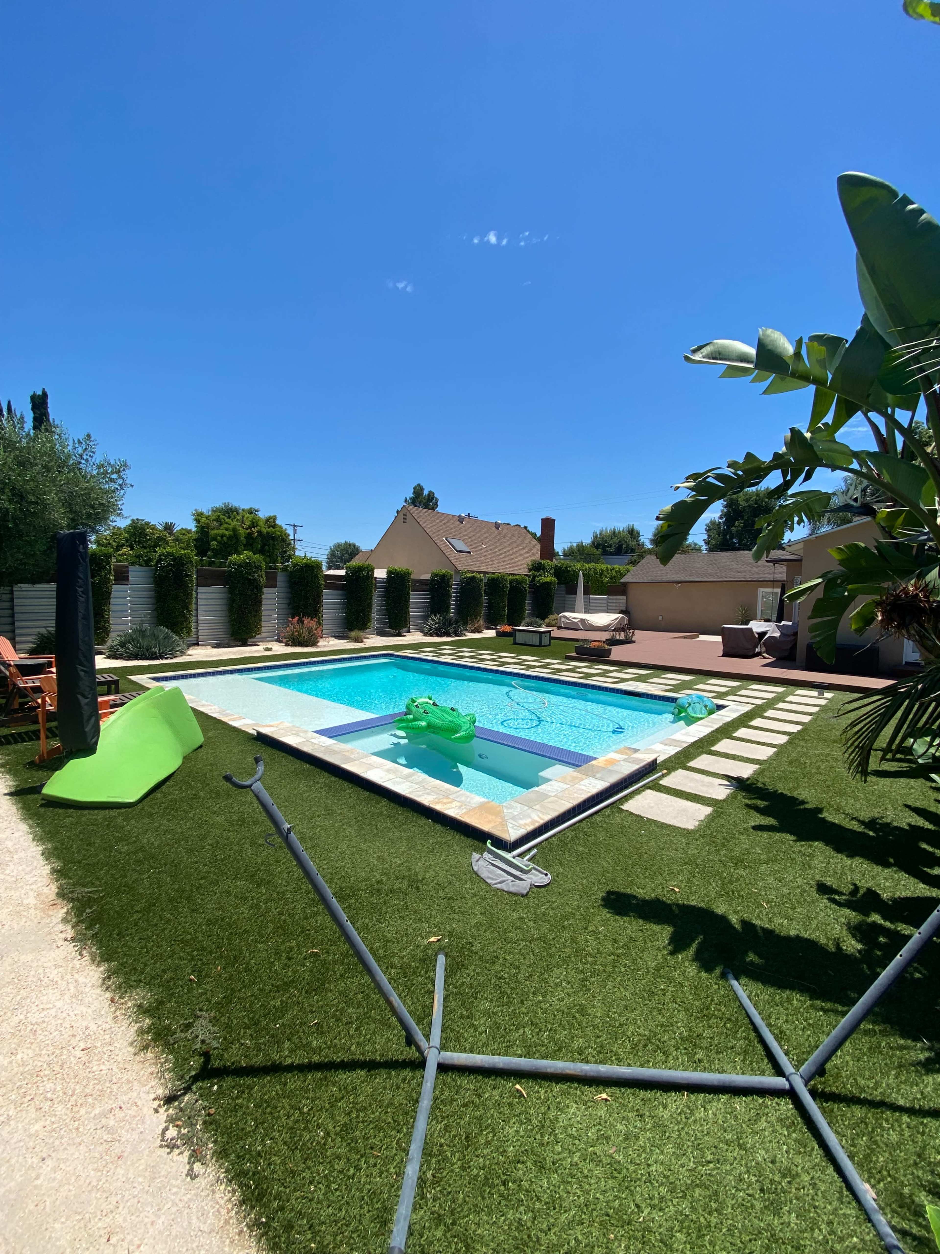 A backyard scene features a swimming pool surrounded by green grass, lounge chairs, and tropical plants under a clear blue sky.