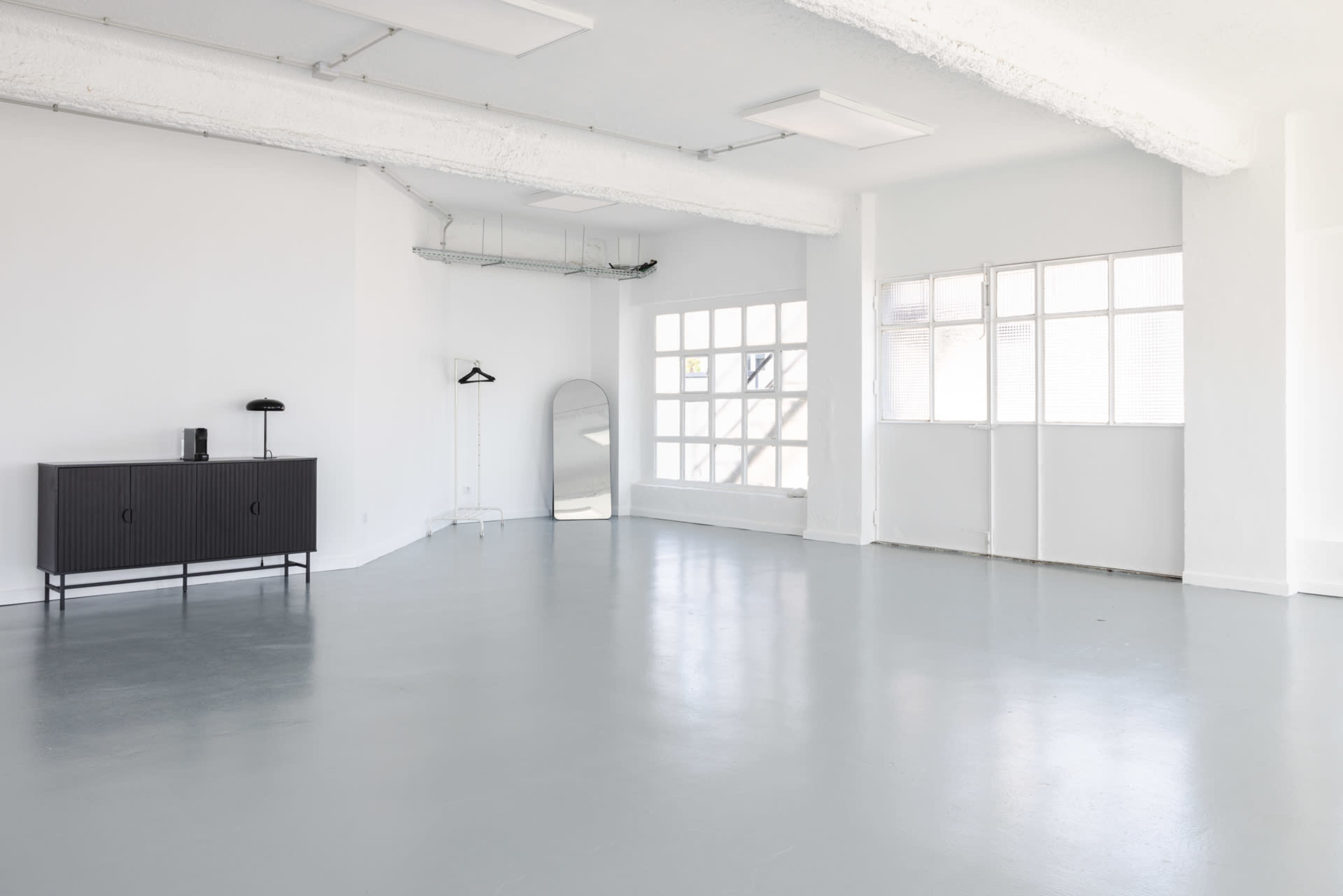 The image shows a bright, minimalistic room with white walls, a polished gray floor, a black cabinet, a mirror, and large windows letting in natural light.
