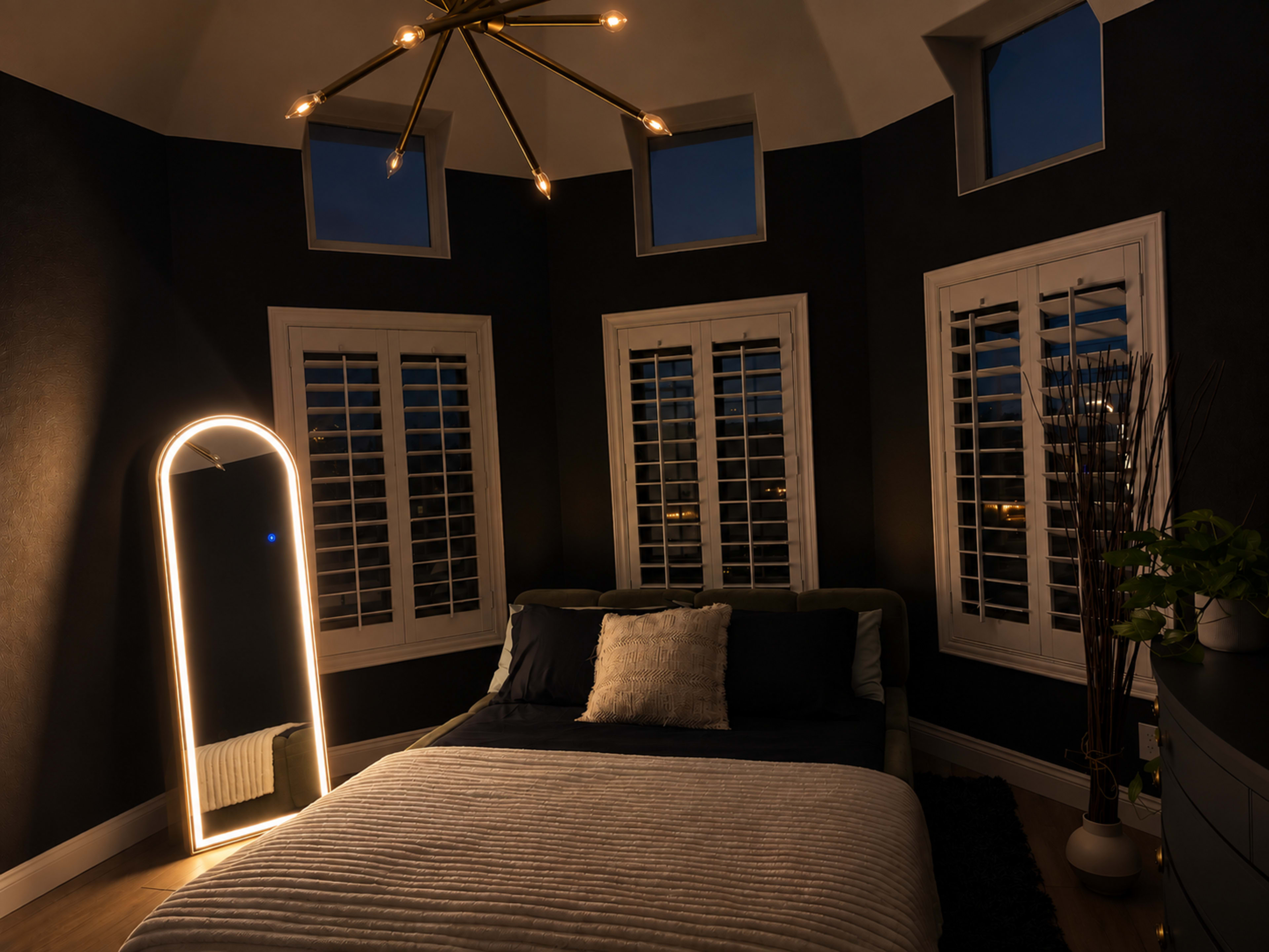 The image shows a modern bedroom with a bed, decorative mirror, and multiple windows, all set against dark walls and illuminated by soft lighting.