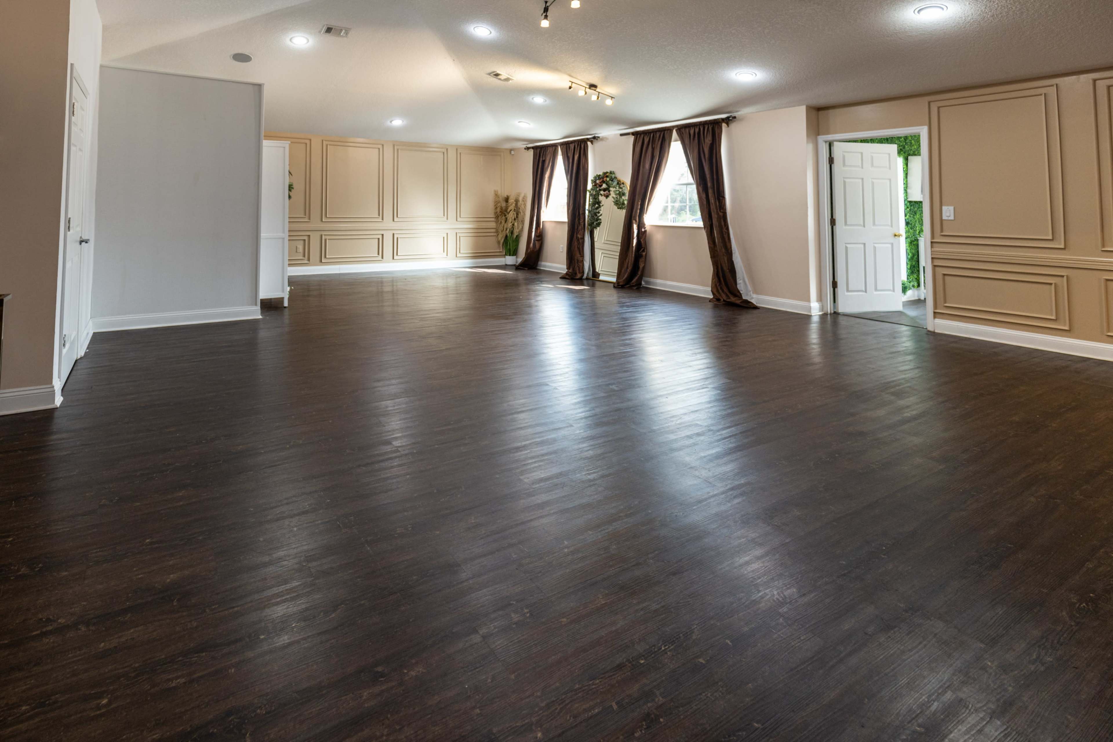 A spacious interior room with dark wood flooring, light-colored walls, and large windows covered by brown curtains.