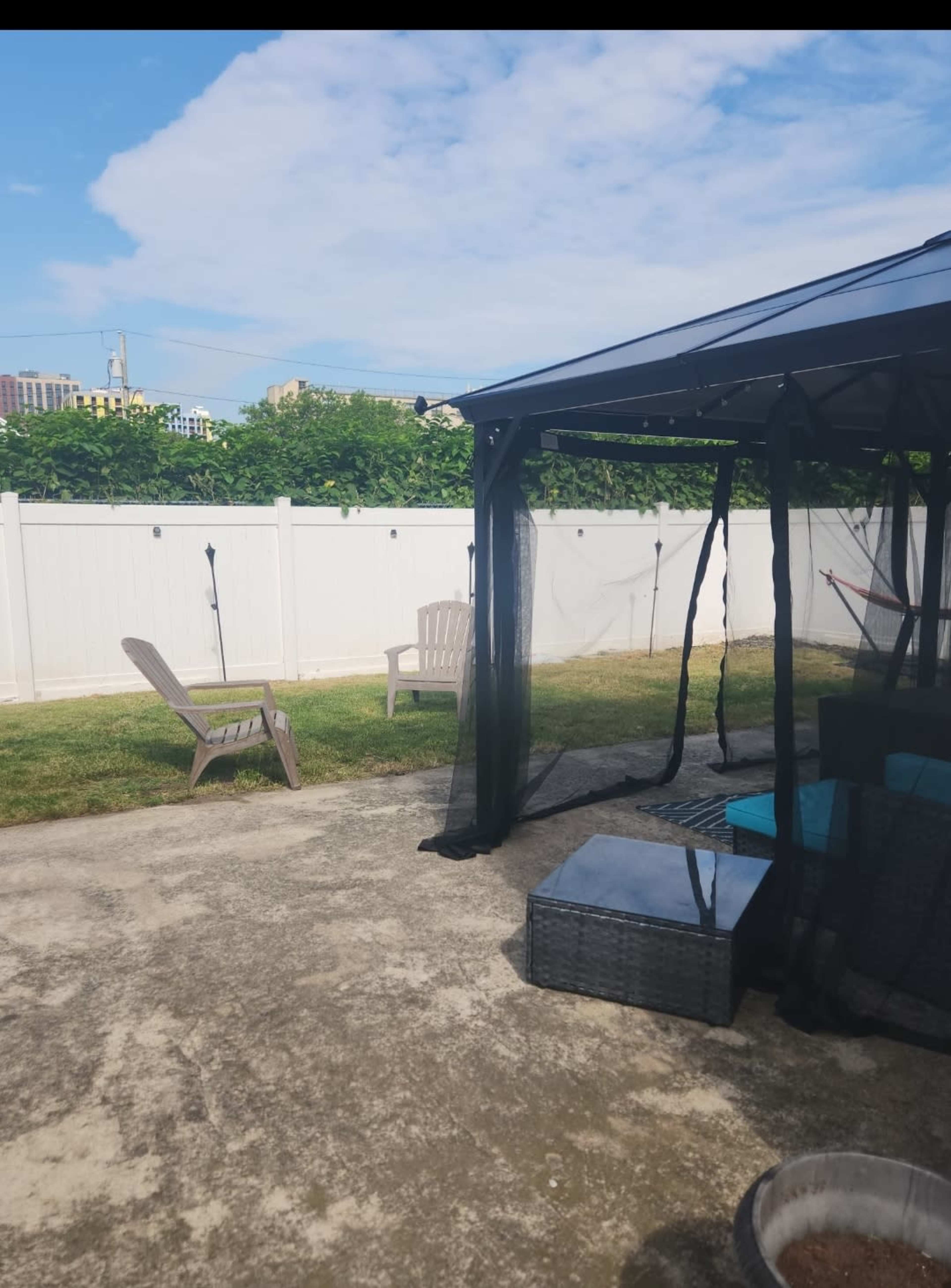 A backyard with a patio, a shaded gazebo, a single chair, and a white fence in the background.