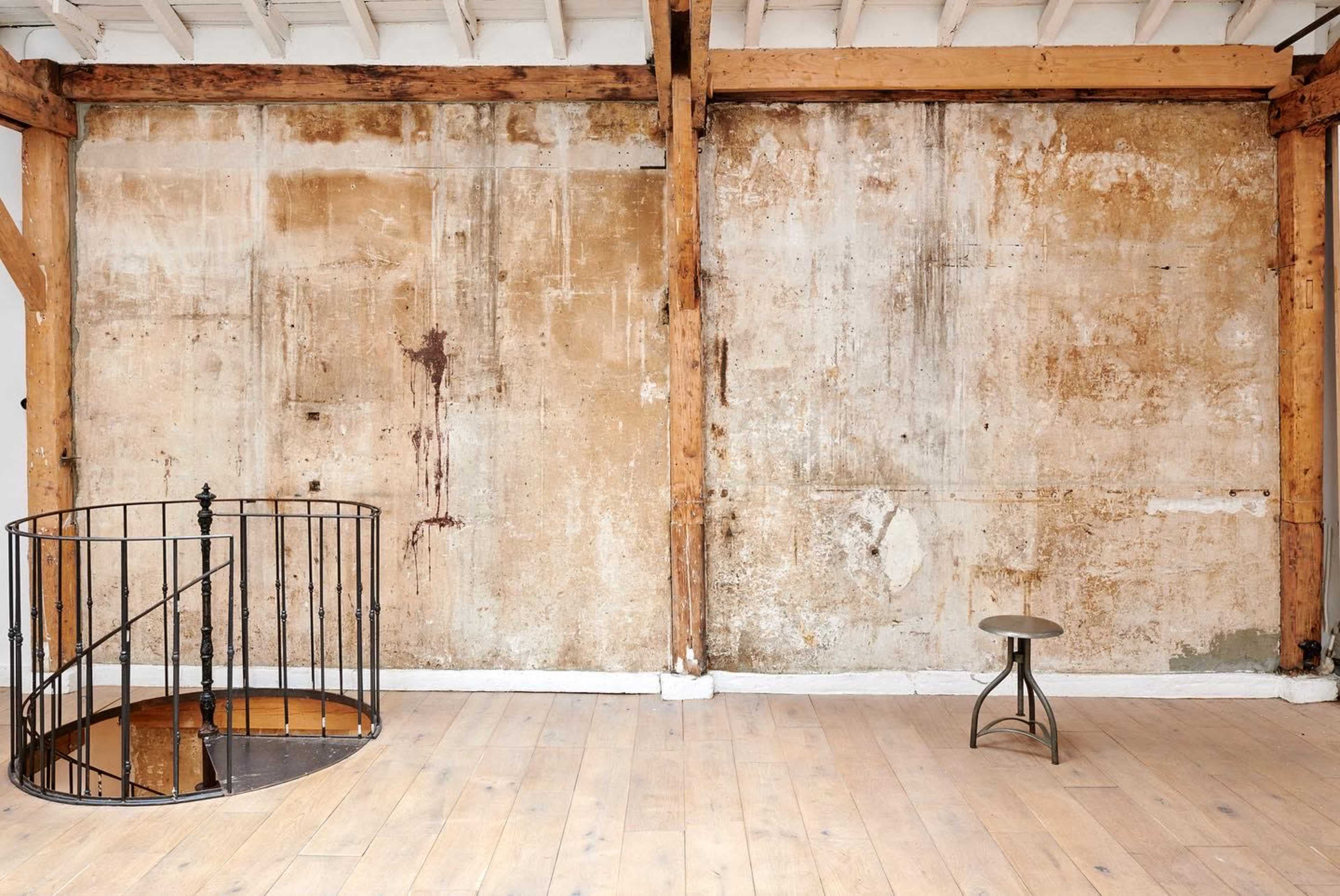 A spacious room with a wooden floor, featuring an exposed wall with peeling paint and wooden beams, along with a circular metal staircase and a small metal stool.