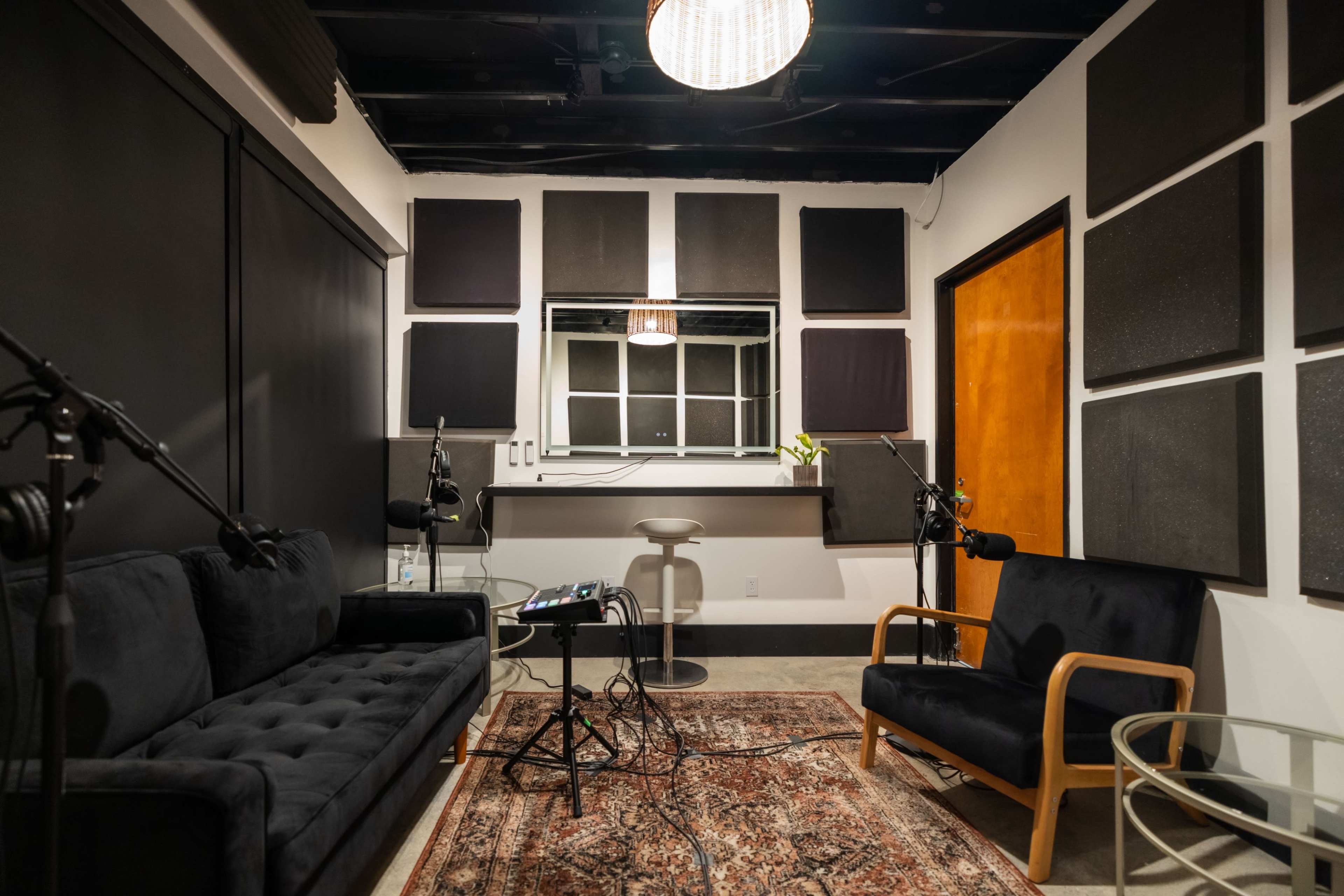 A recording studio featuring a dark sofa and chair, a mixing console on the floor, soundproof wall panels, and a large window behind a countertop.