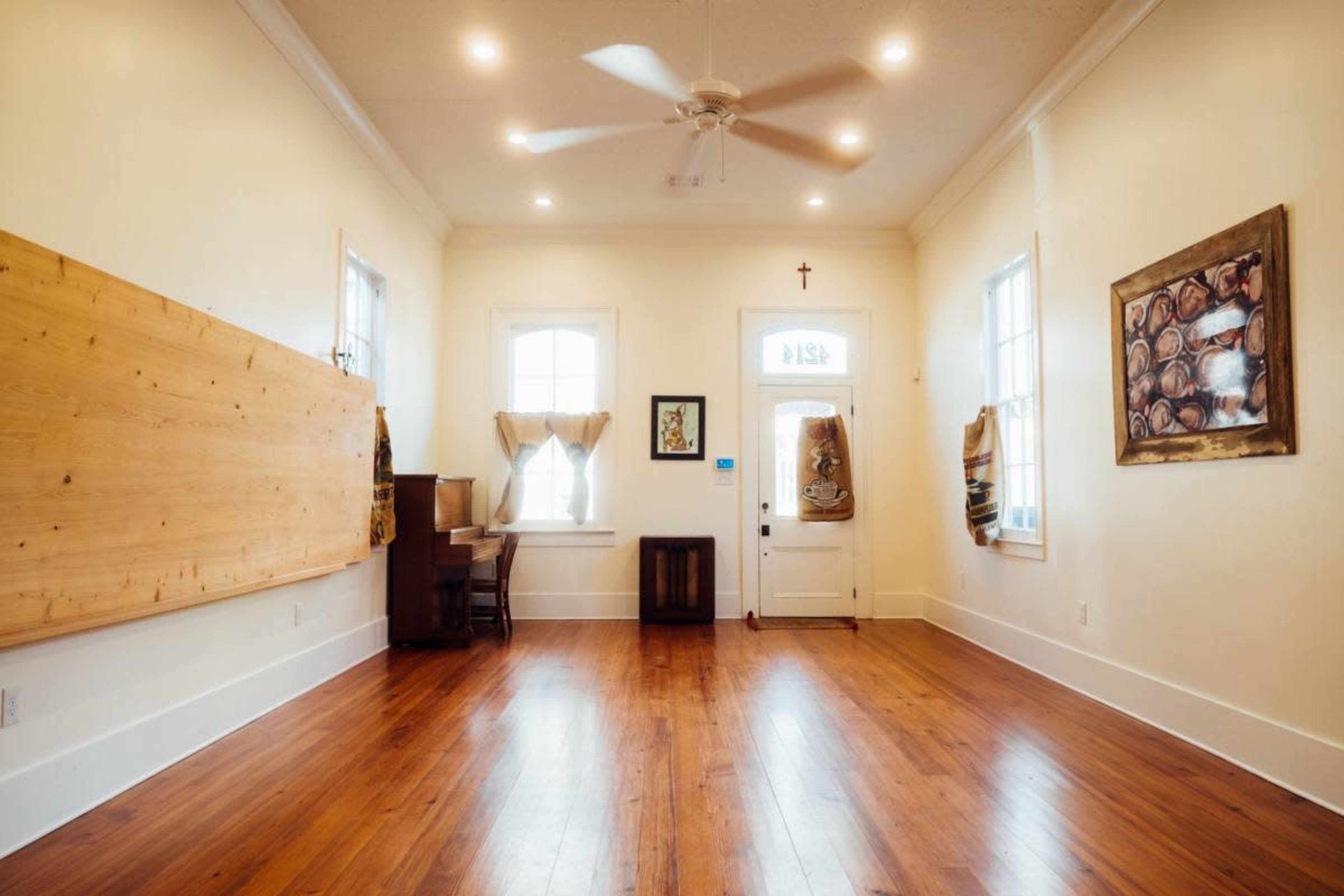 The interior of a spacious, bright room features wooden flooring, a piano against one wall, and large windows with light curtains.