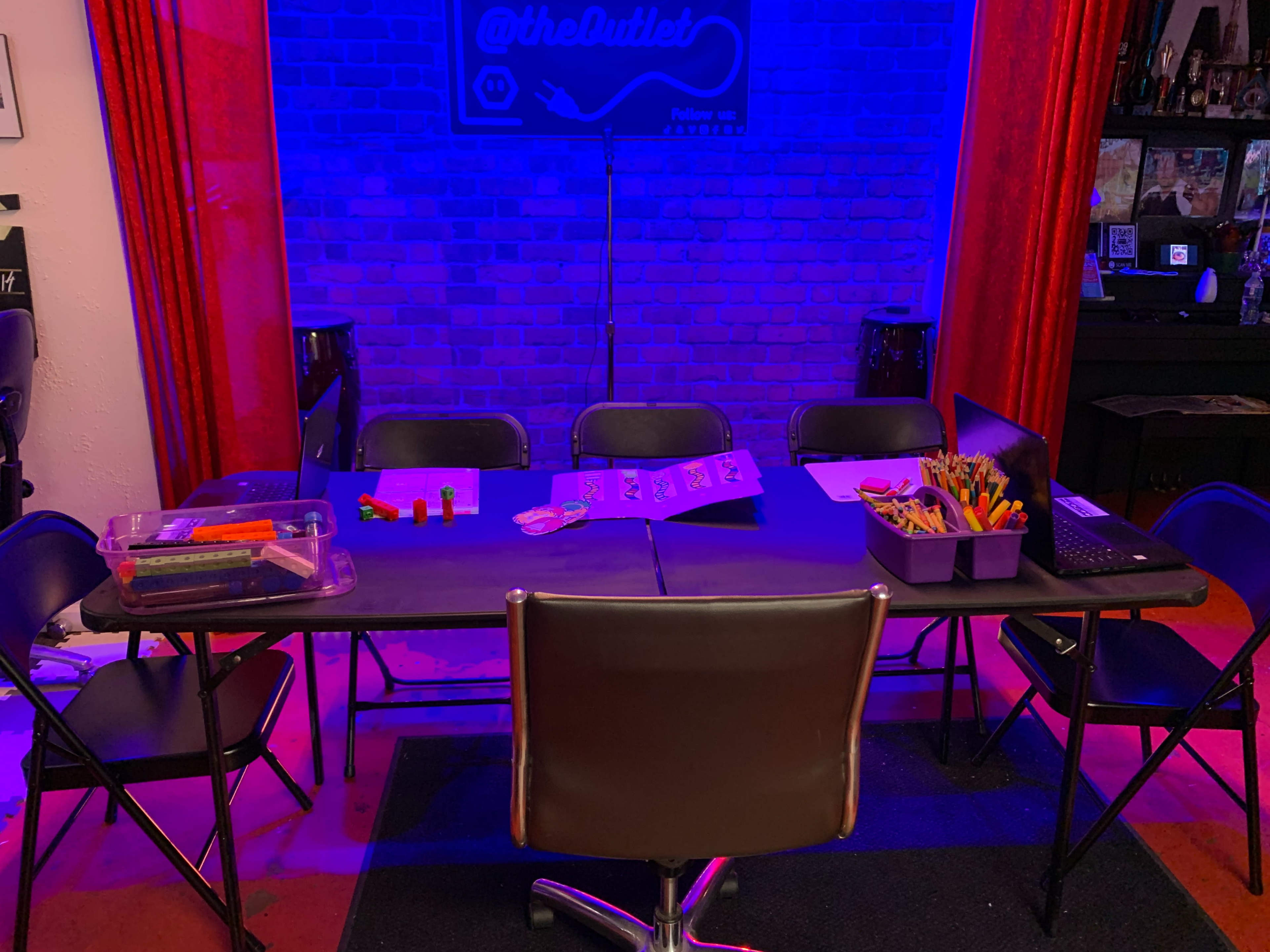 A rectangular table with various art supplies and a laptop is set up in a room, surrounded by several black chairs under blue and red lighting.