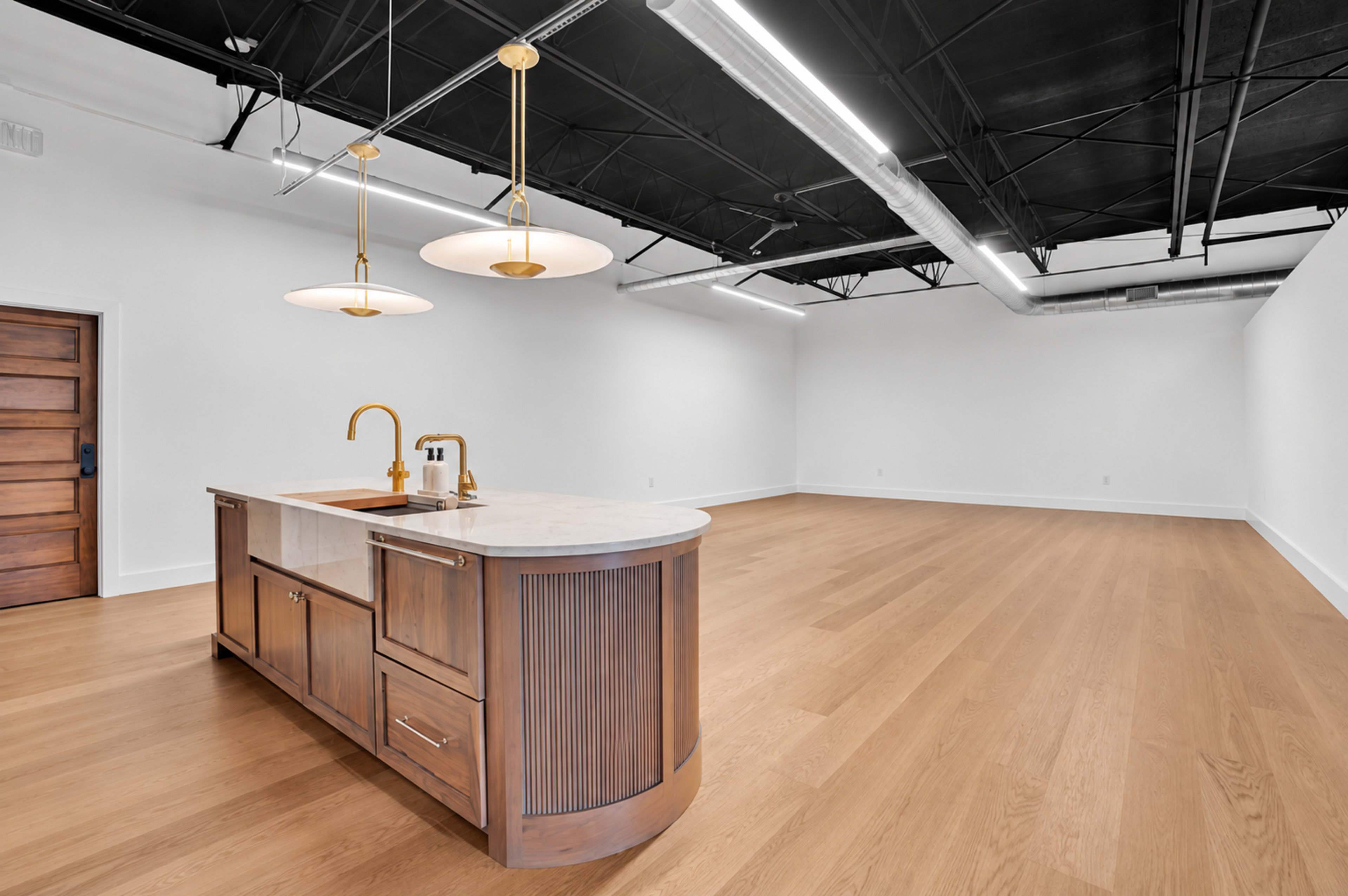 A modern kitchen island with a marble countertop and gold fixtures is positioned in a large, empty room with hardwood flooring and black ceiling beams.