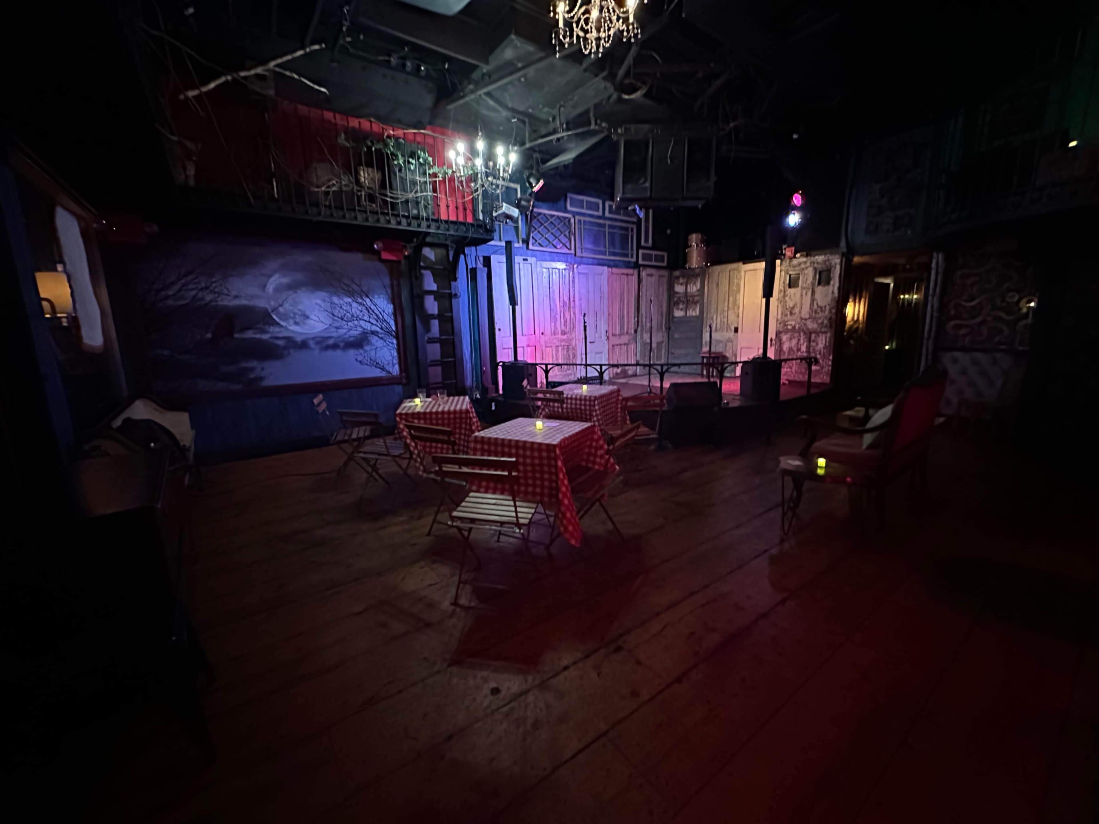 The image shows an empty, dimly lit venue with tables covered in red and white checkered cloths and a backdrop of colorful lighting.